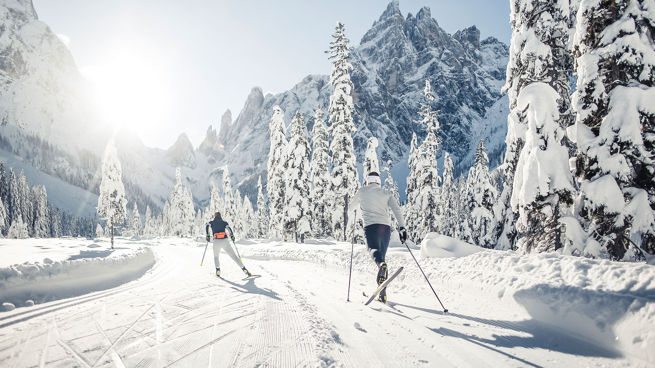 Two people cross-country skiing on a snowy mountain trail surrounded by snow-covered trees and peaks.