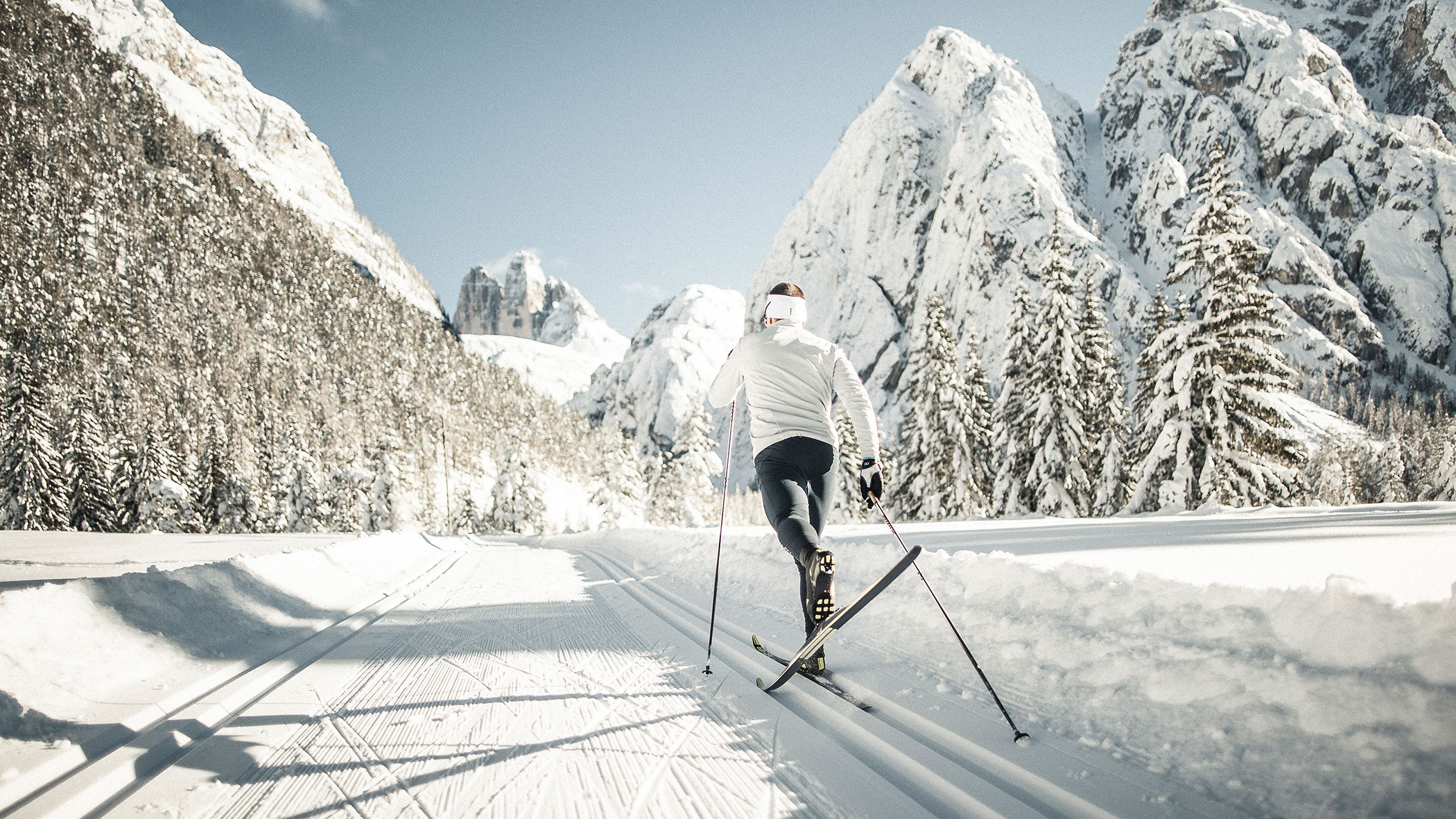 A person cross-country skiing on a snowy trail surrounded by tall mountains and pine trees.