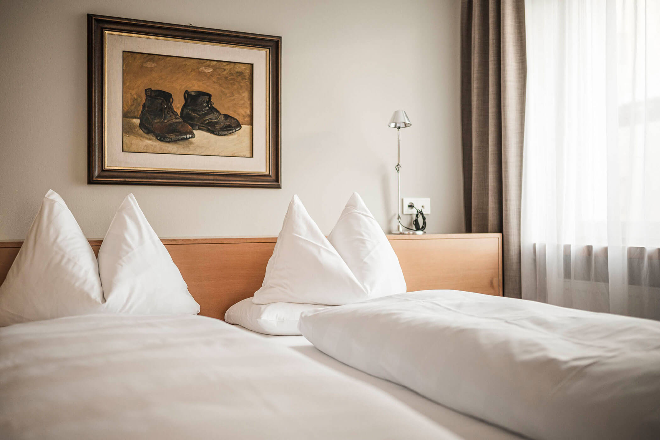 Two neatly made single beds with white linens beneath a painting of boots in a cozy, sunlit room.