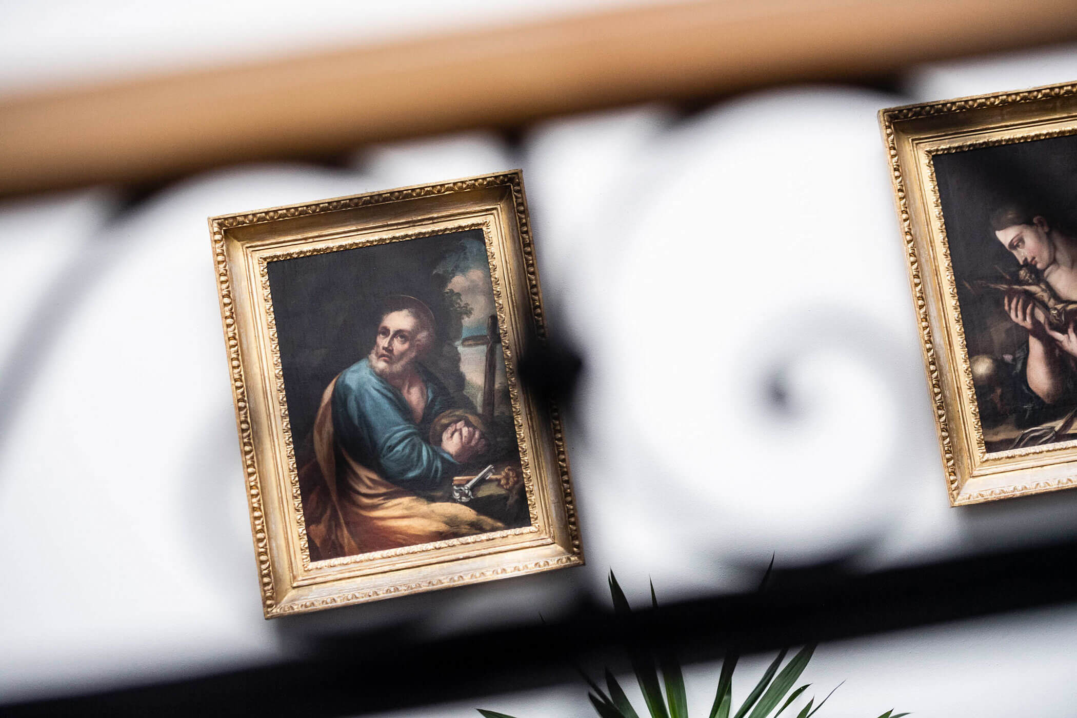 Two framed classical paintings on a white wall, partially obscured by a black decorative railing.