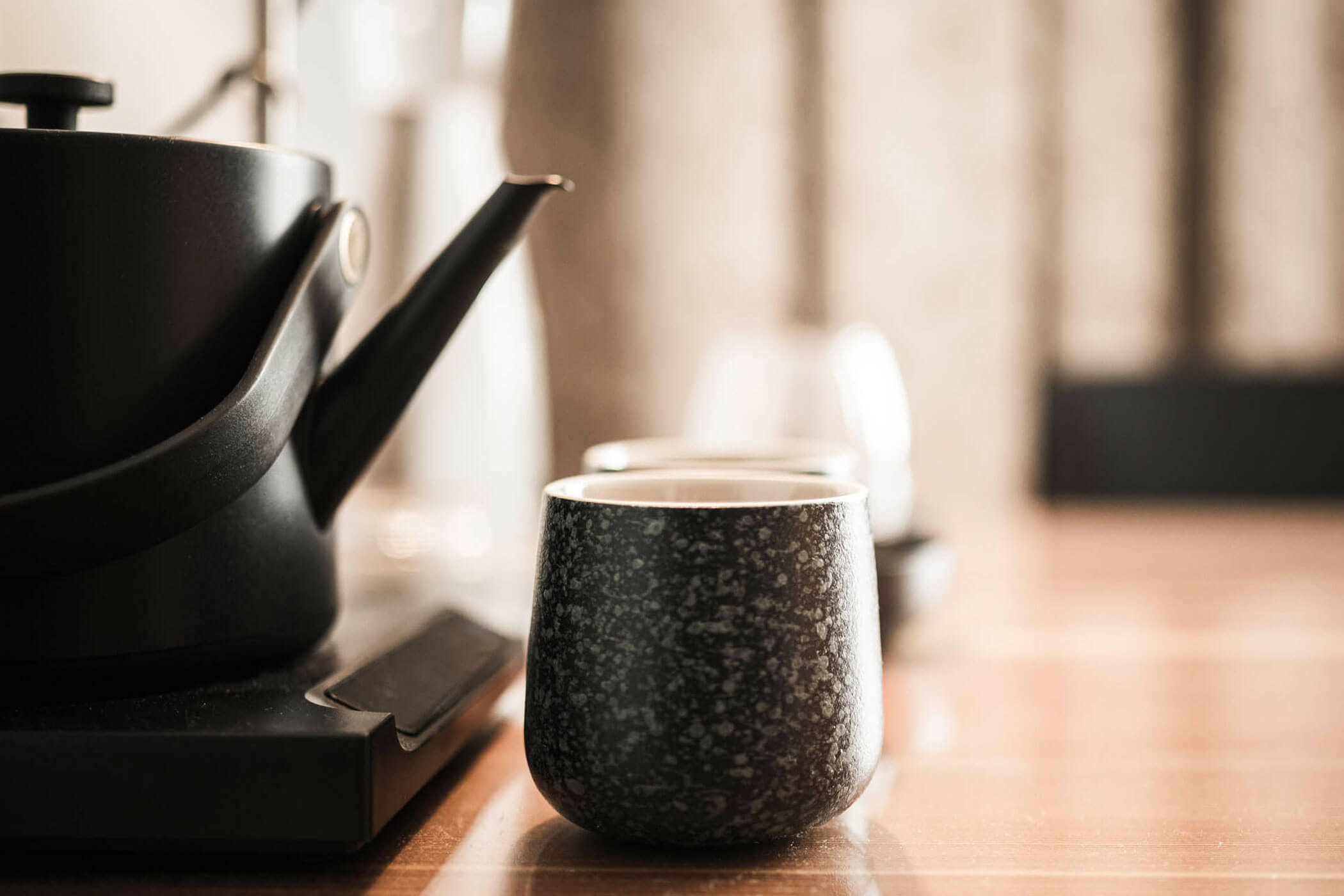 A black teapot and a speckled ceramic cup sit on a wooden table in soft, warm lighting.