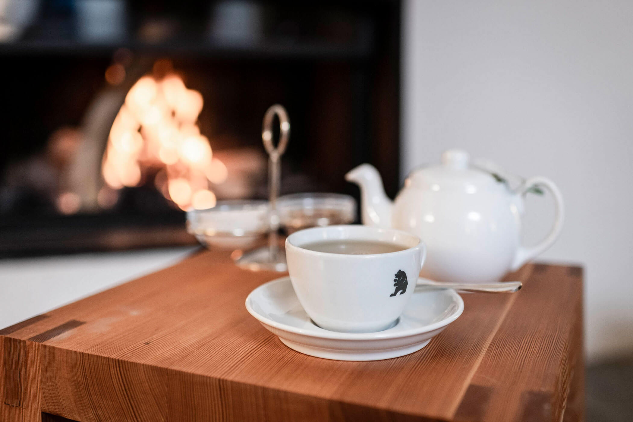A cup of tea on a wooden table with a teapot and a fireplace burning in the background.