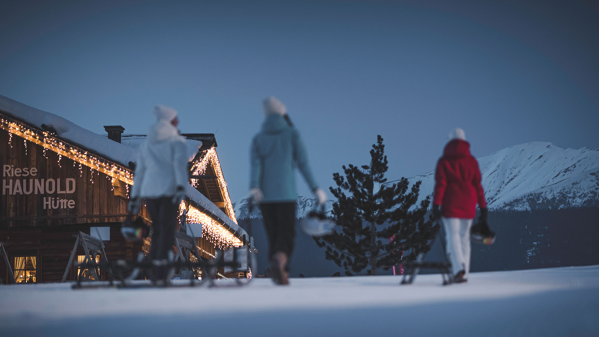 Three people walk in snow near a lit cabin at dusk, with mountains in the background.