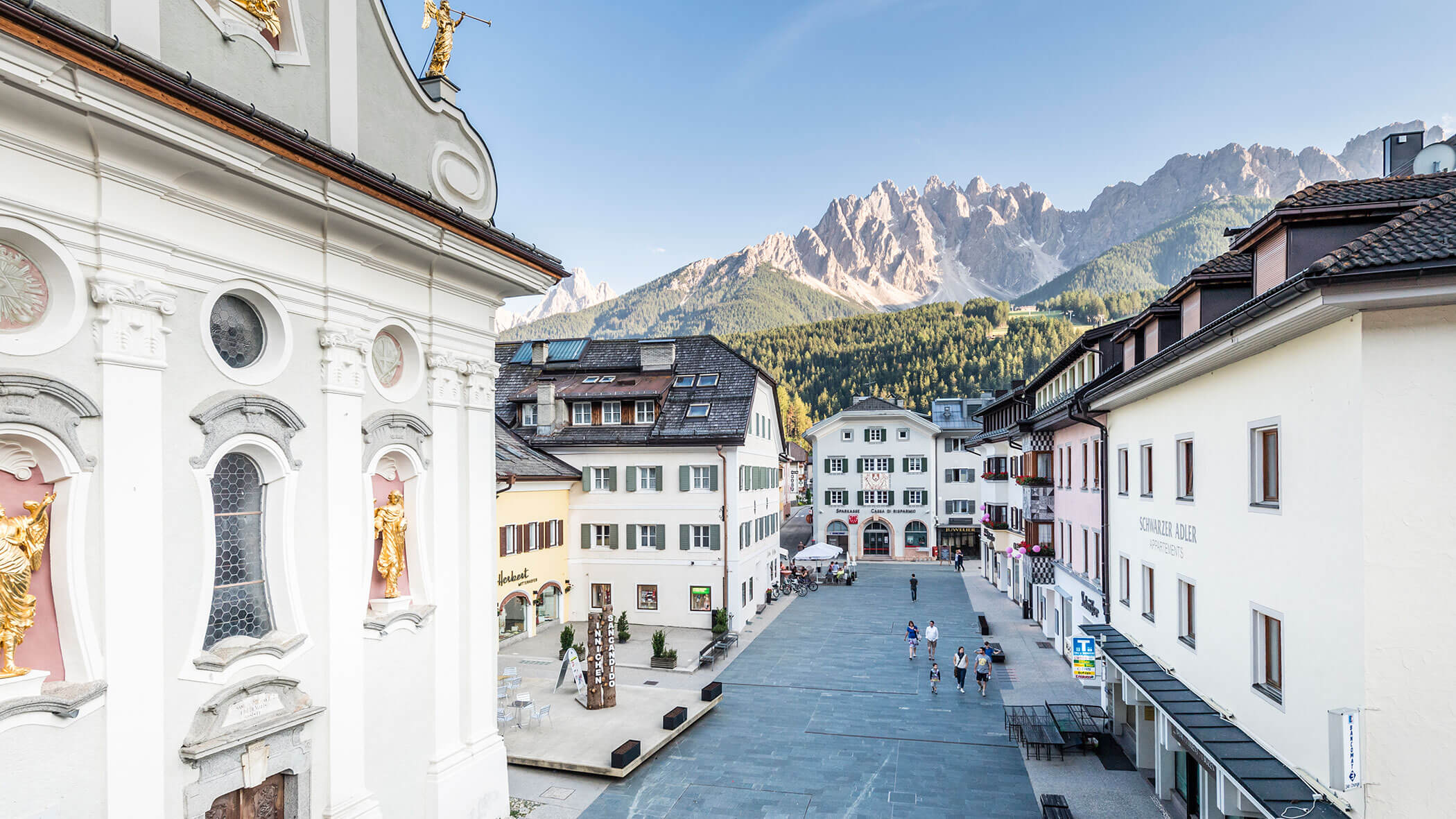 Una pittoresca piazza di città europea con edifici alpini e montagne sullo sfondo, sotto un cielo limpido.