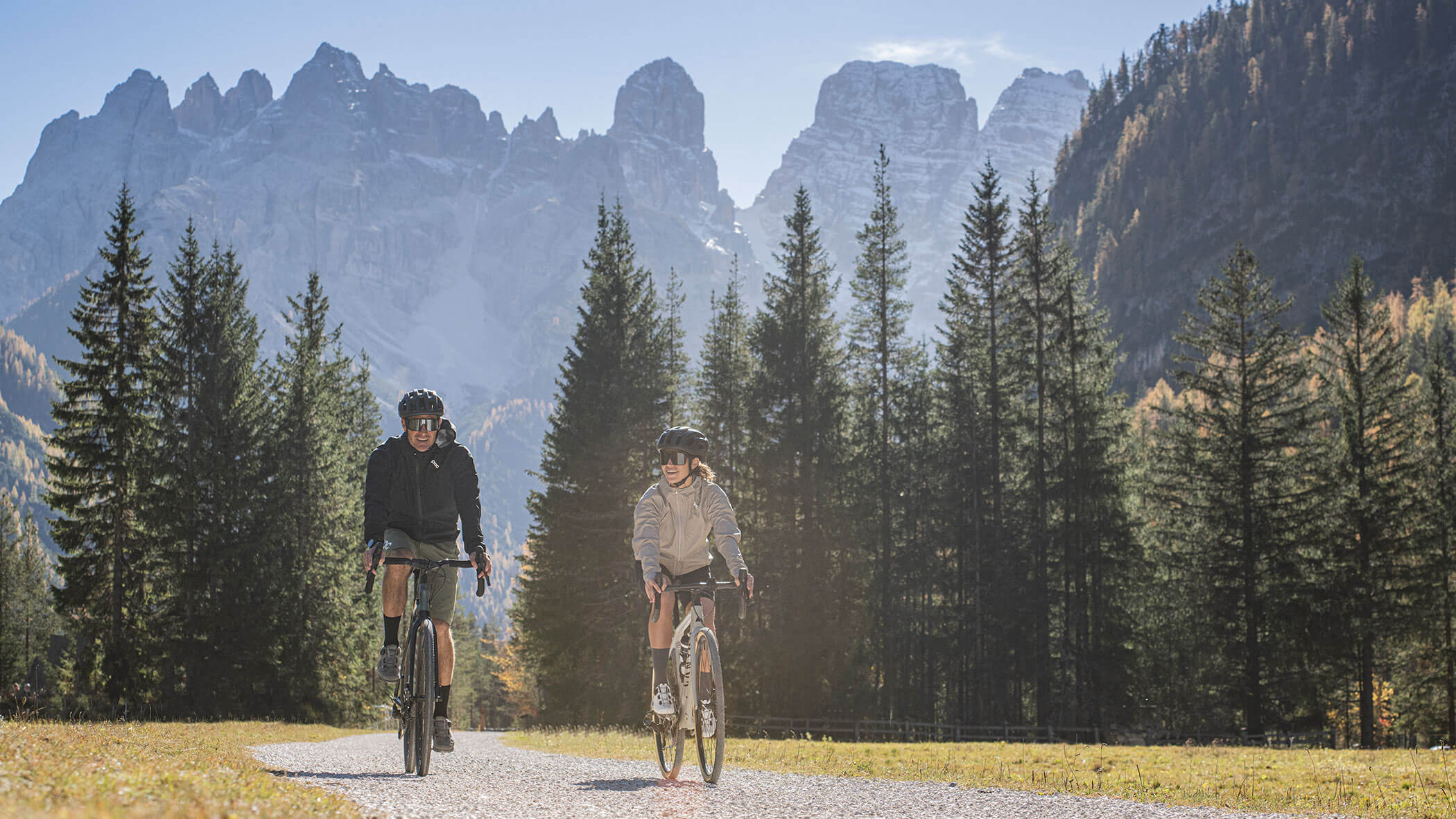 Two people ride bicycles on a path through a forested valley with tall mountains in the background.