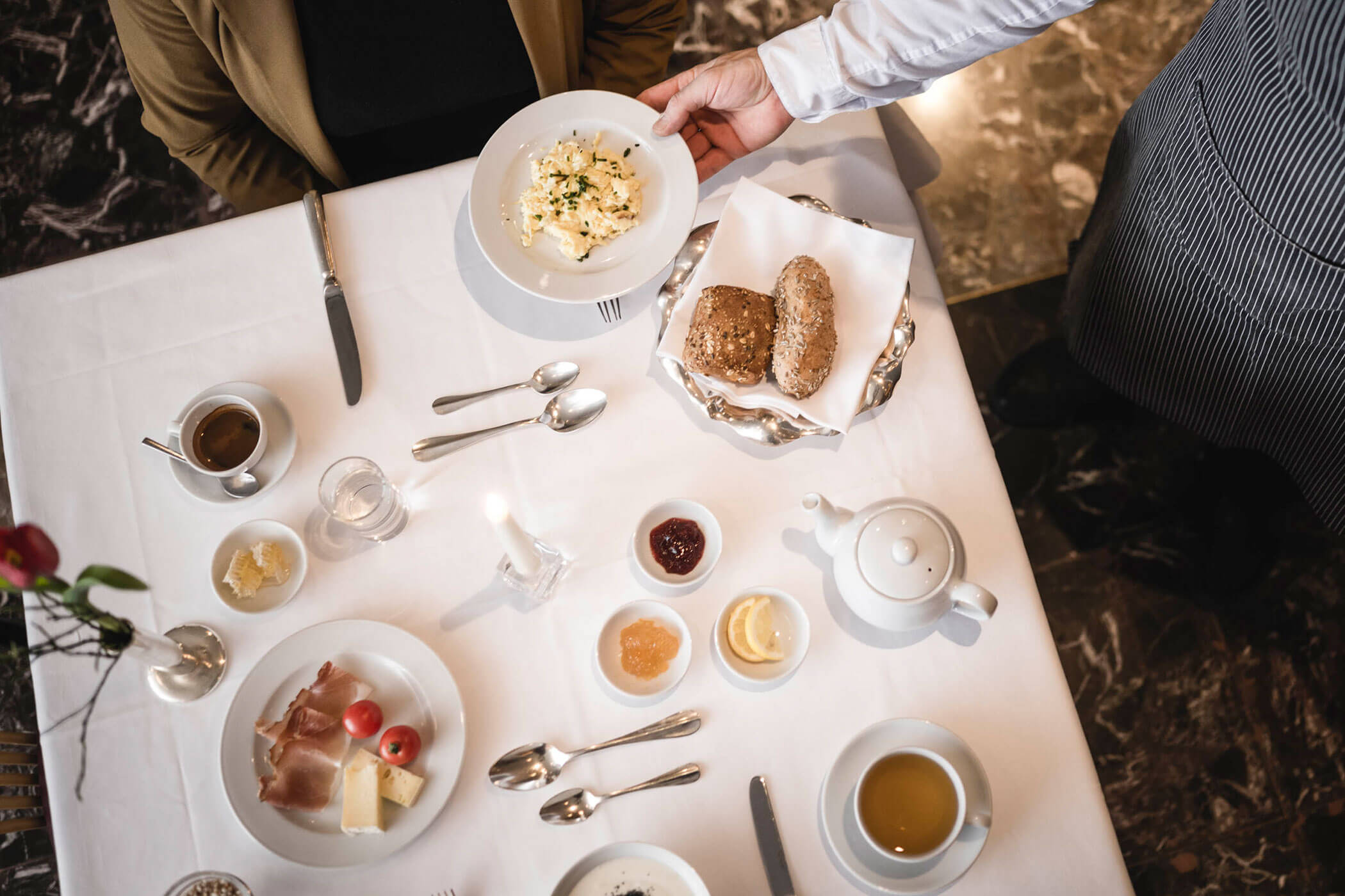 A waiter serves scrambled eggs at a breakfast table set with bread, spreads, tea, and cold cuts.