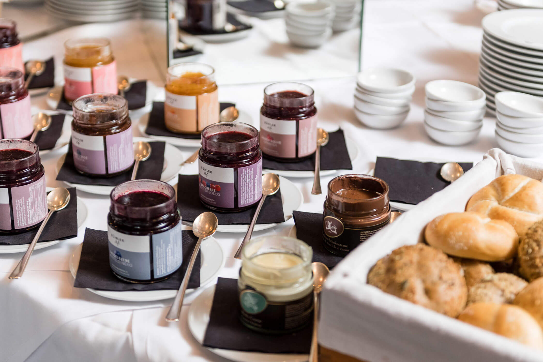 Jars of spreads and jams with spoons, next to a basket of assorted bread rolls on a table.