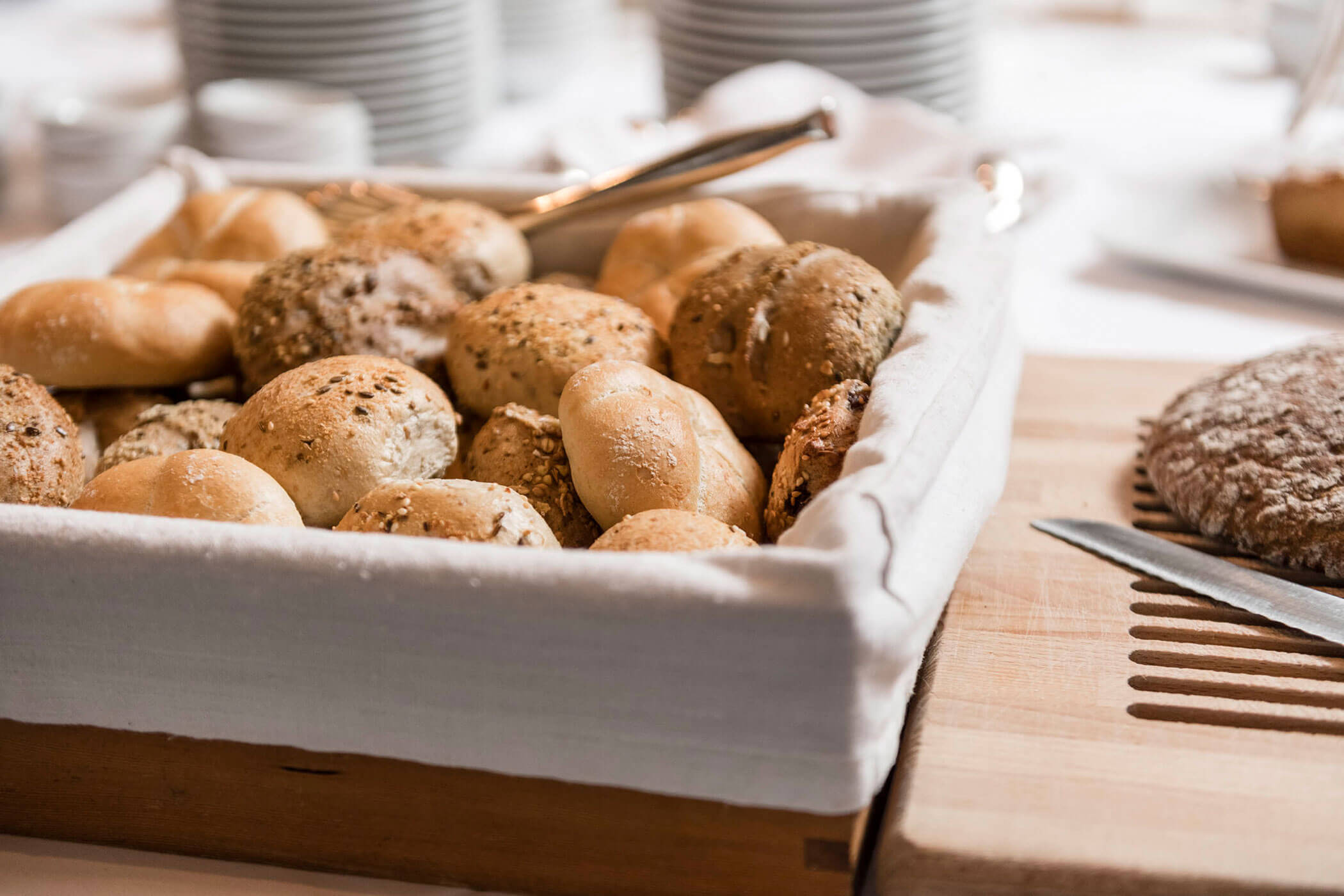 A basket of assorted bread rolls and a loaf of bread on a cutting board with a knife.