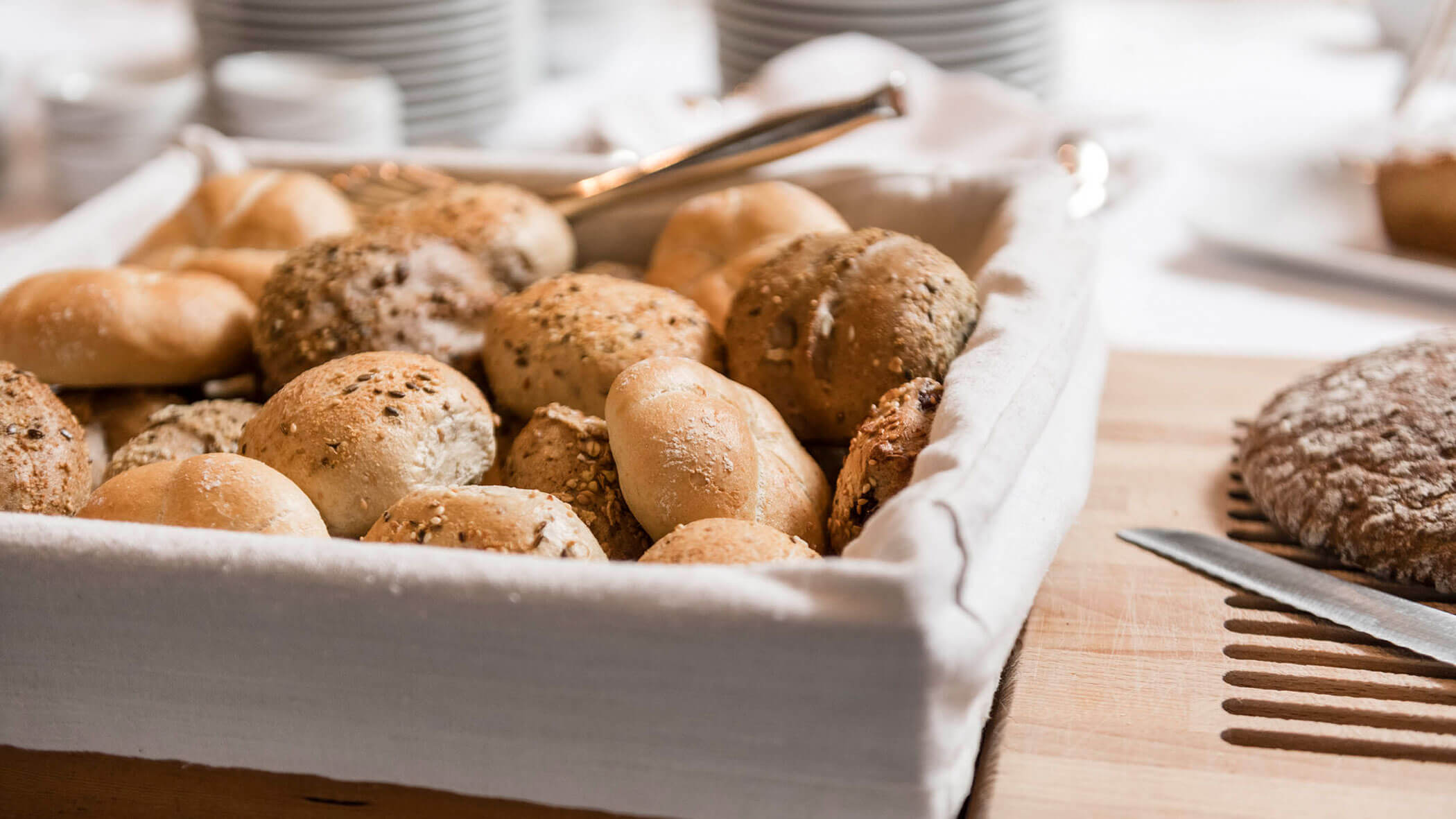 A basket of assorted bread rolls on a table with a knife and sliced bread nearby.