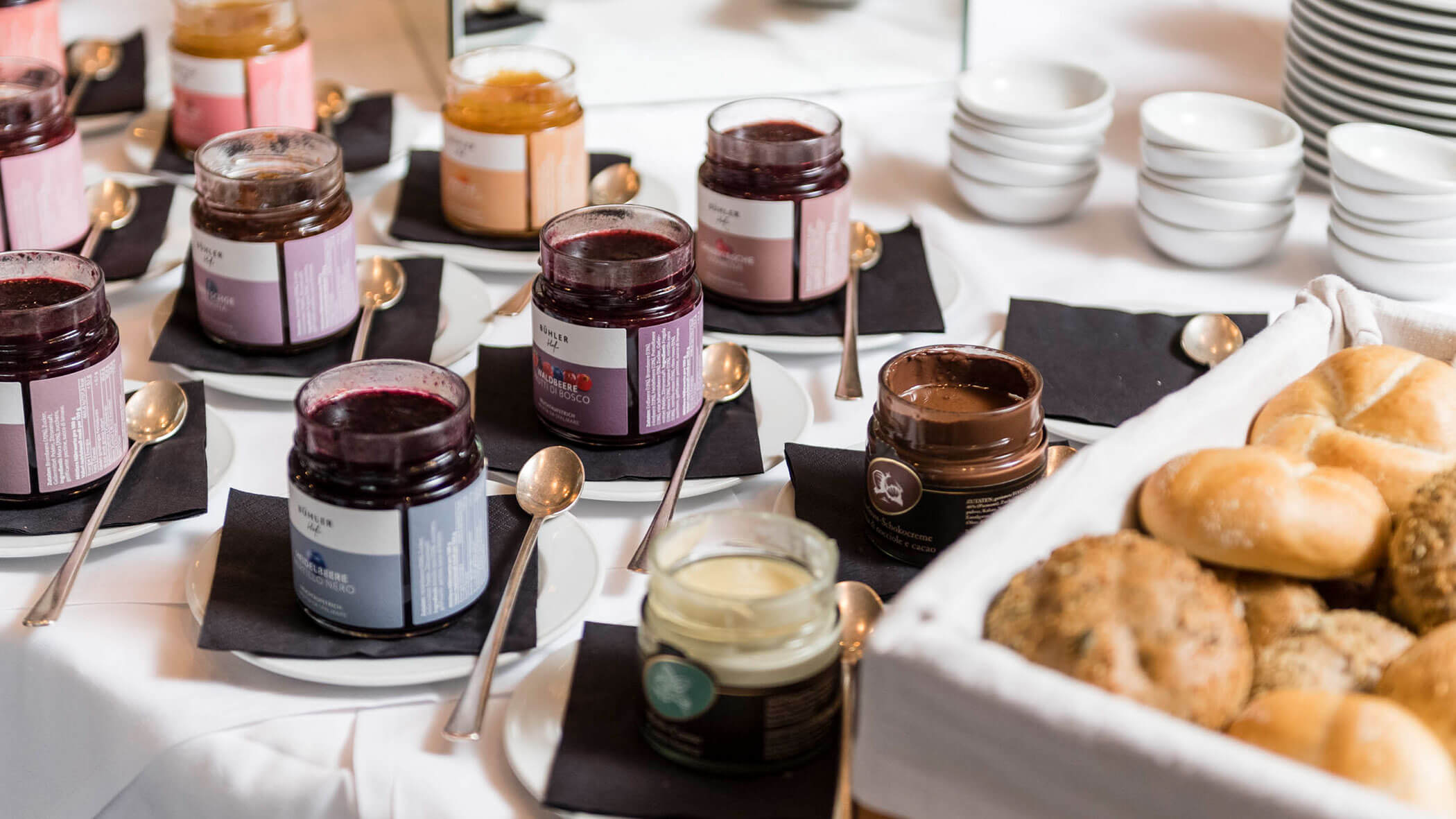 Jars of jam with spoons and a basket of bread rolls on a white tablecloth, with stacked bowls in the background.