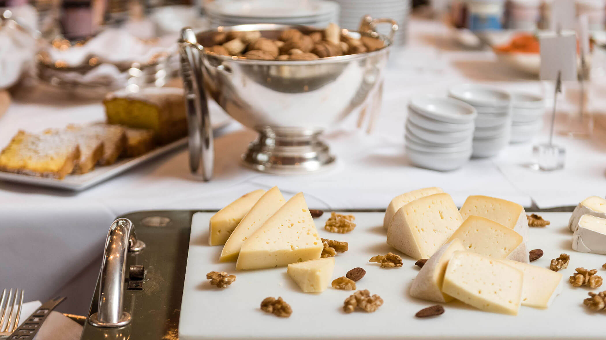 Assorted cheese wedges with walnuts and almonds on a platter, with bowls and dishes in the background.