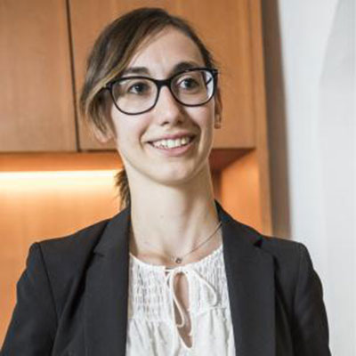 Woman with glasses and brown hair in a ponytail, wearing a black blazer and white blouse, smiling indoors.