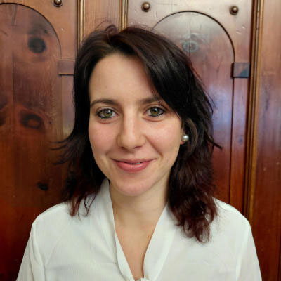A woman with dark hair and a white blouse smiles in front of wooden doors.