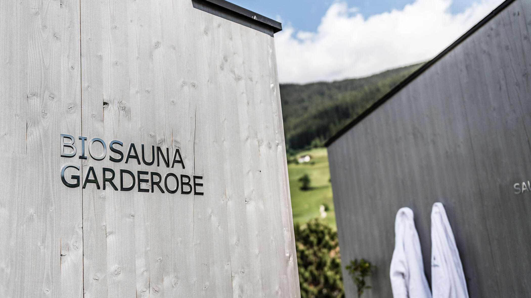 Sign reads Biosauna Garderobe on a wooden wall; robes hang outside against a mountain landscape.