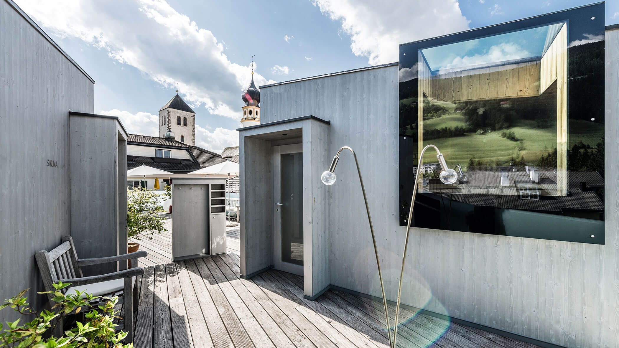 Modern rooftop terrace with wooden decking, minimalist seating, and a scenic view of a church tower and countryside.