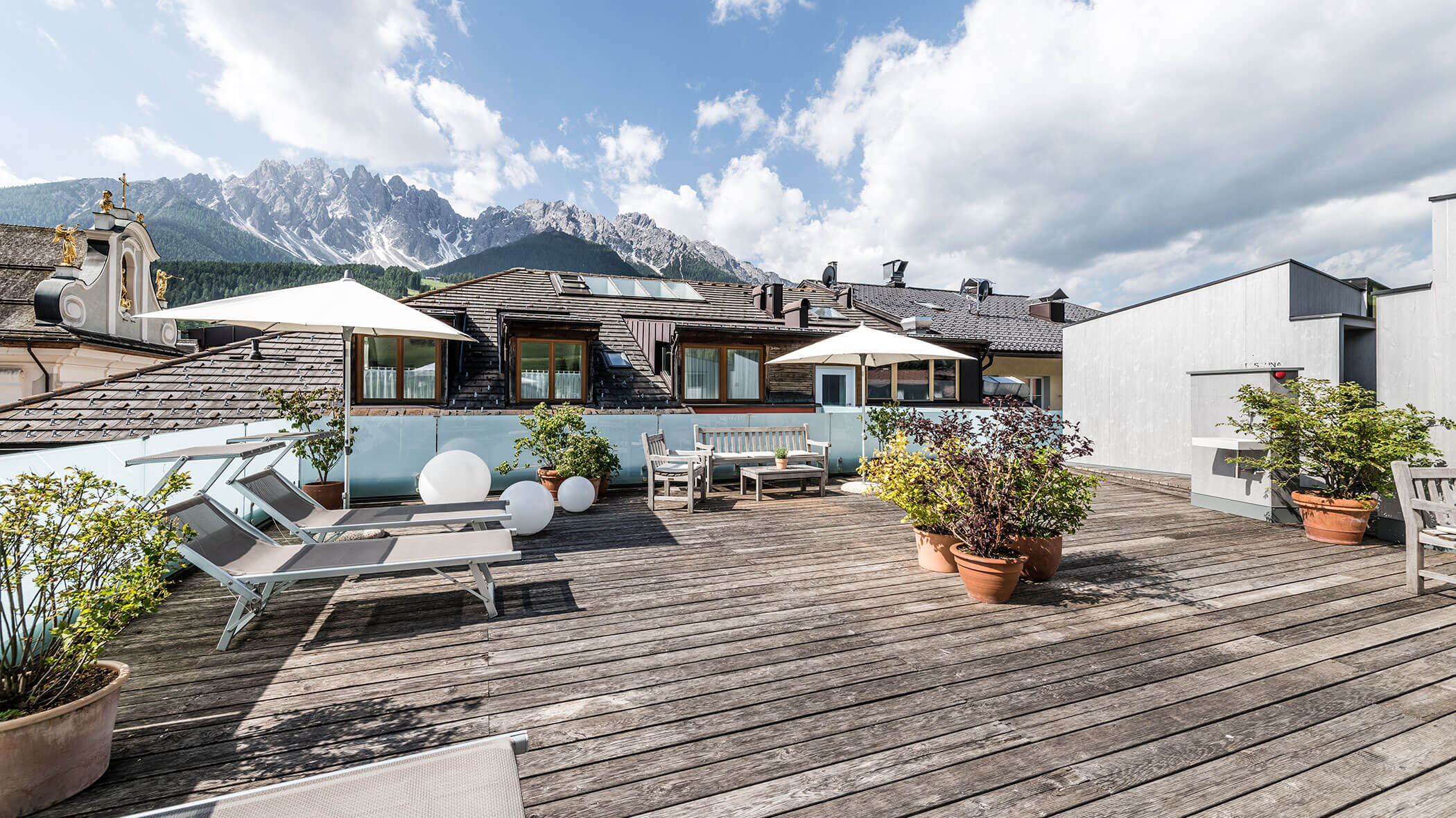 Rooftop terrace with lounge chairs, umbrellas, potted plants, and mountain views under a partly cloudy sky.