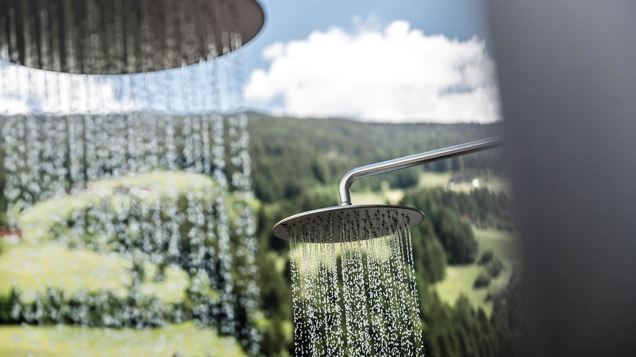 Outdoor showers with water running, set against a scenic background of green hills and a partly cloudy sky.