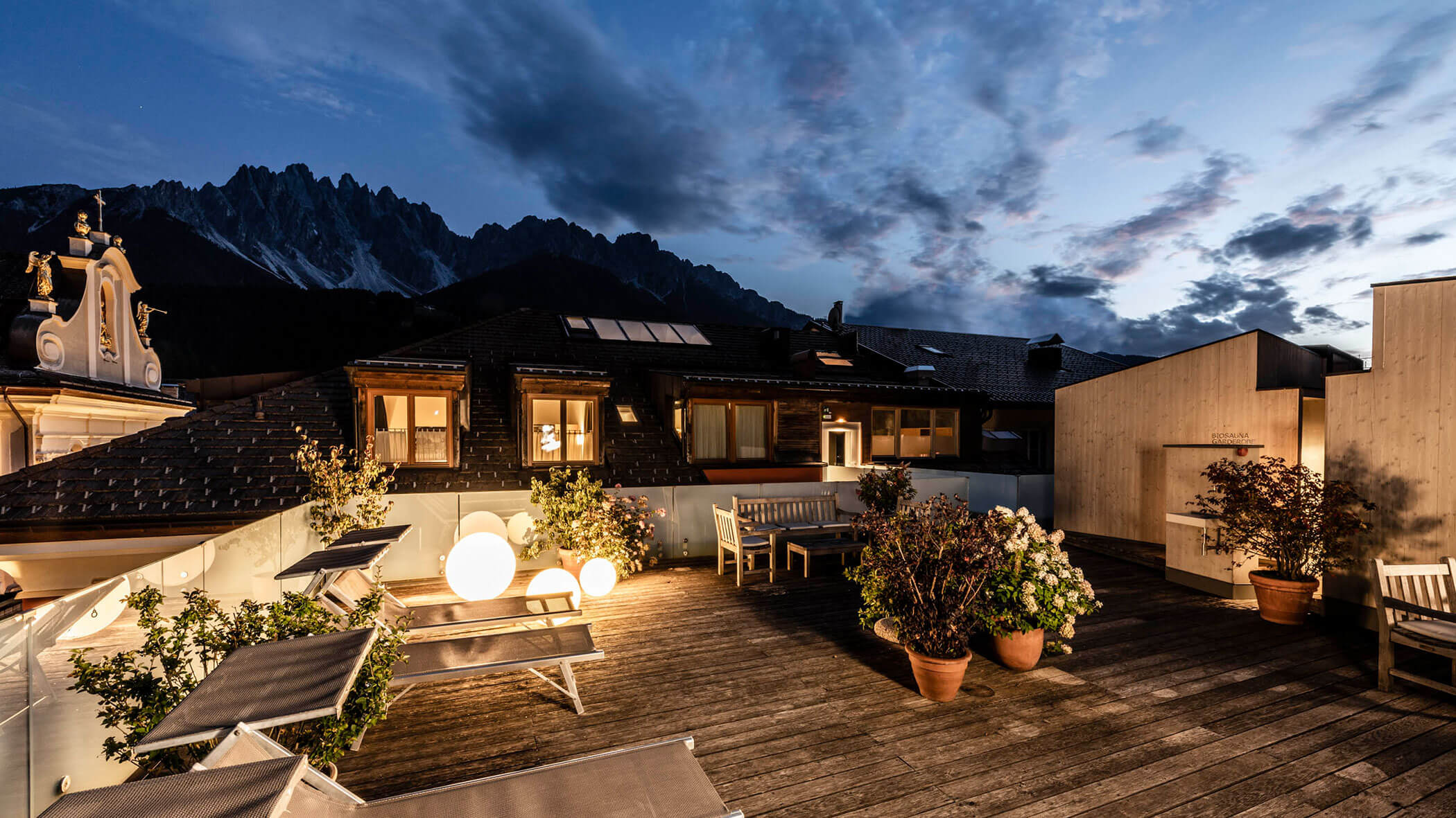 A rooftop terrace at dusk with lounge chairs, potted plants, and glowing round lights, mountains in the background.