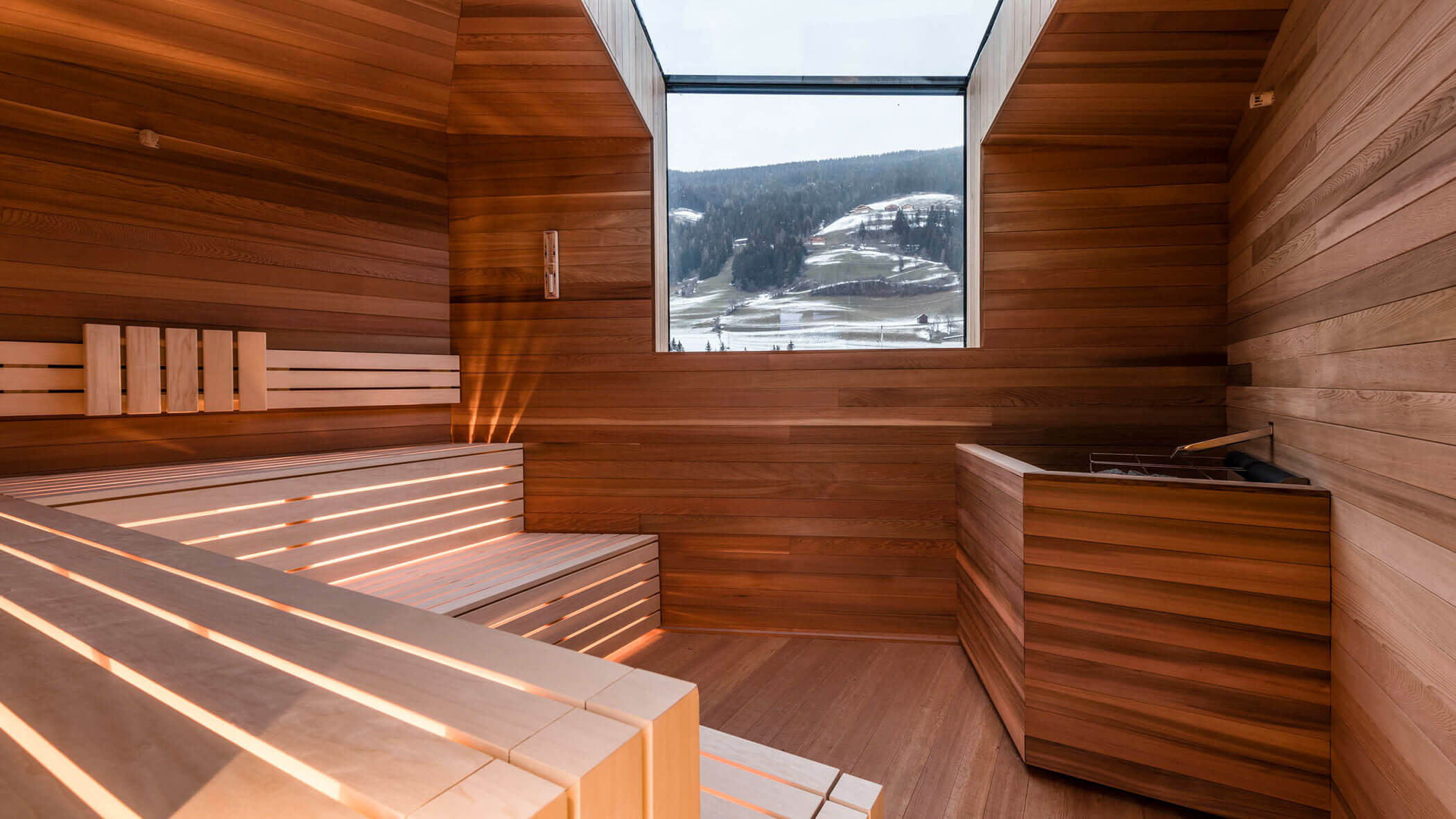 Modern wooden sauna with tiered benches and a large window overlooking a snowy mountain landscape.