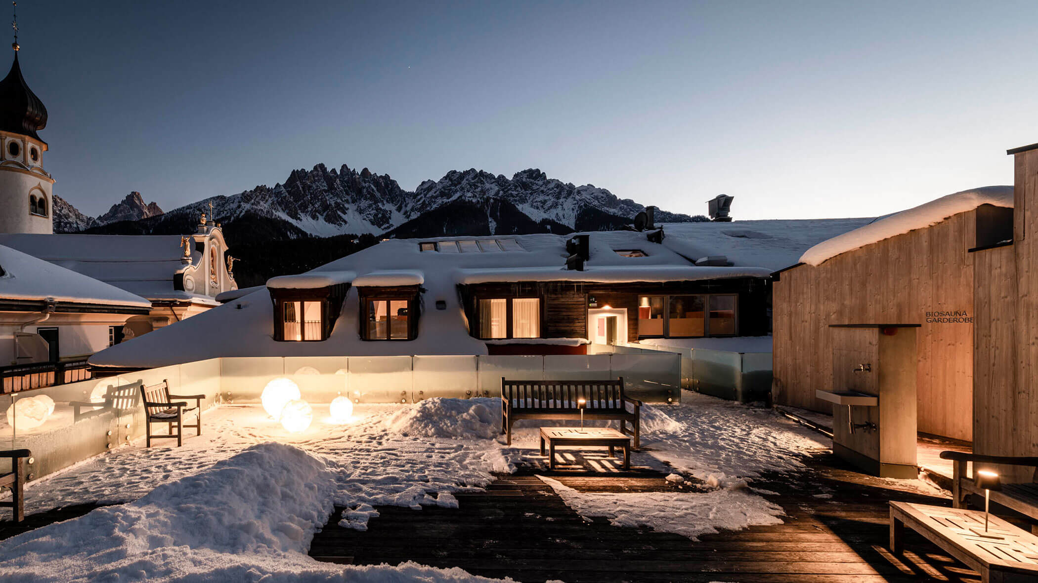 Snow-covered rooftop terrace with benches, glowing lights, and mountain peaks in the background at dusk.