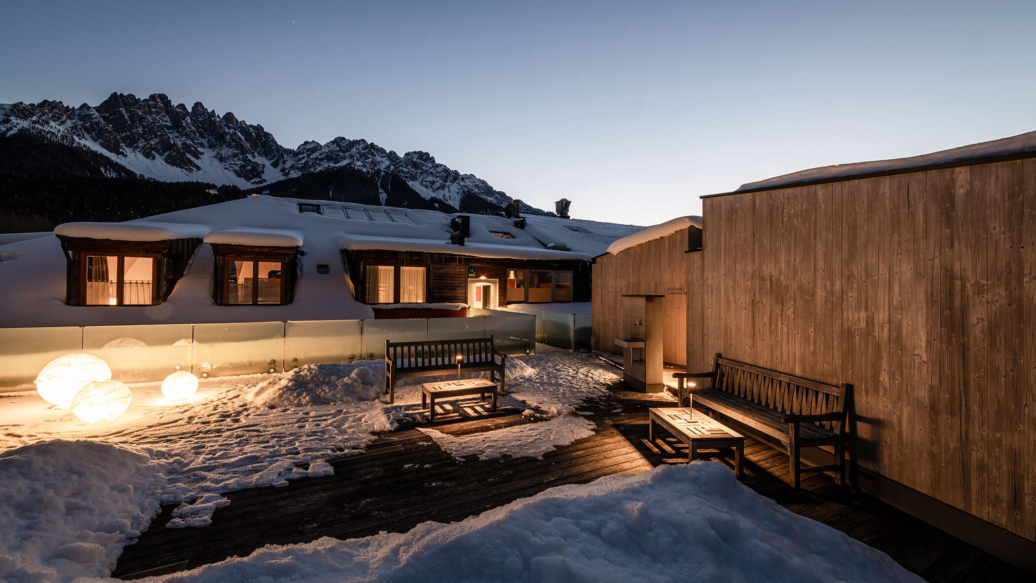 Snow-covered patio with benches, glowing lights, and wooden buildings, set against mountain peaks at dusk.