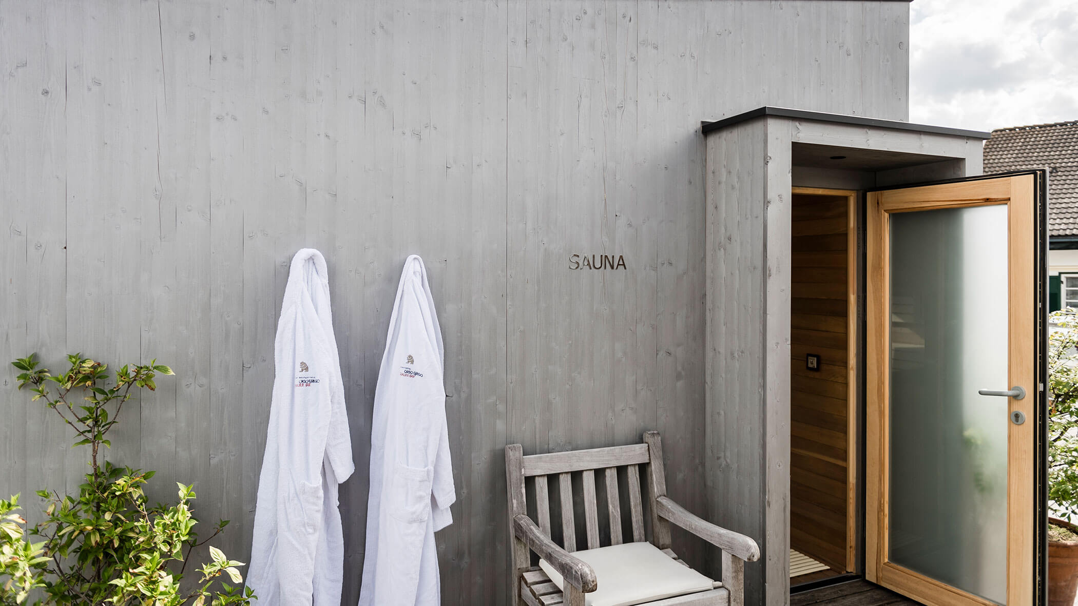Two white robes hang on a gray wooden wall beside a sauna entrance and a wooden chair outdoors.