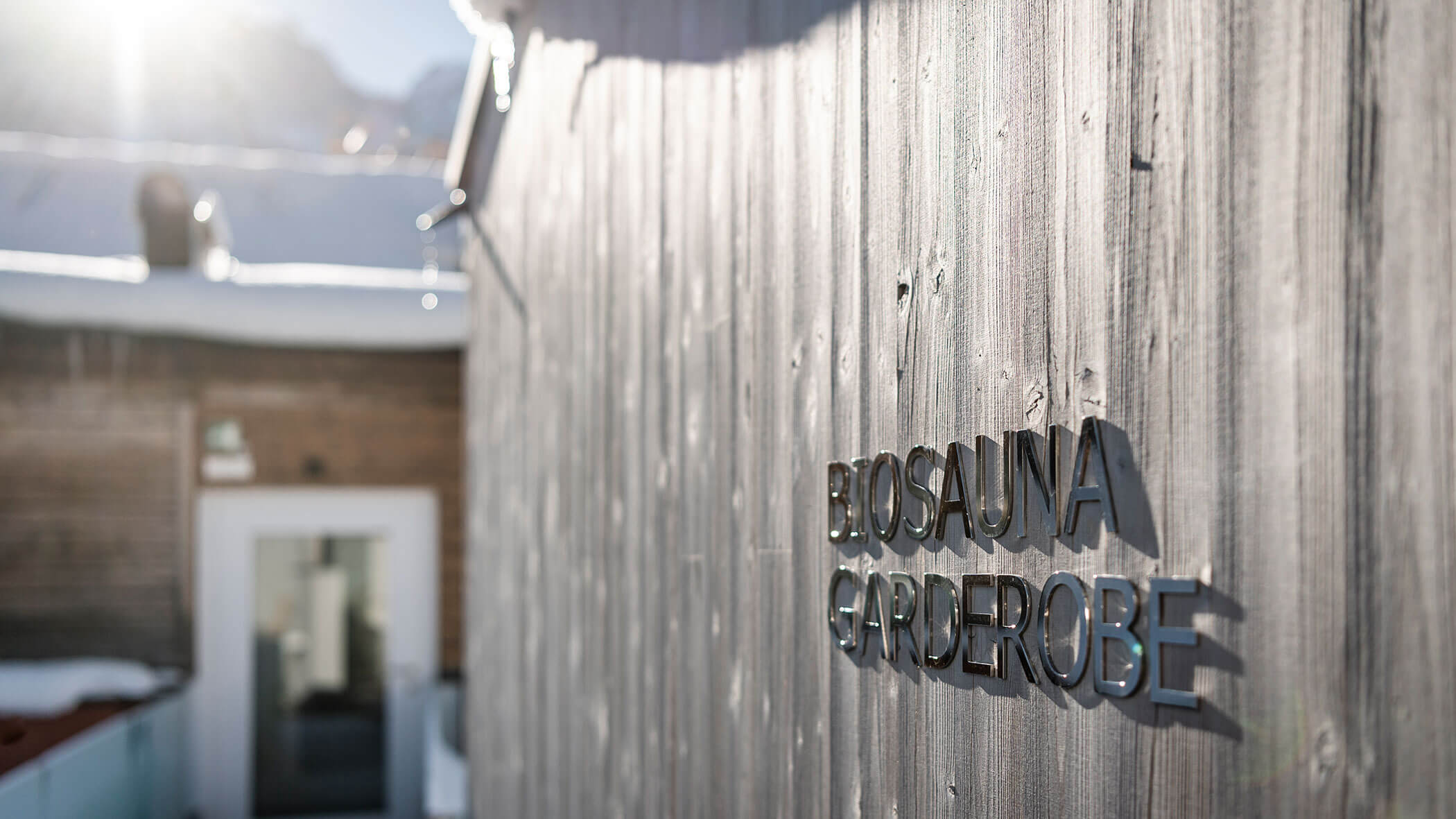 Close-up of a wooden wall with Biosauna Garderobe sign, a building and snowy roof in the background.