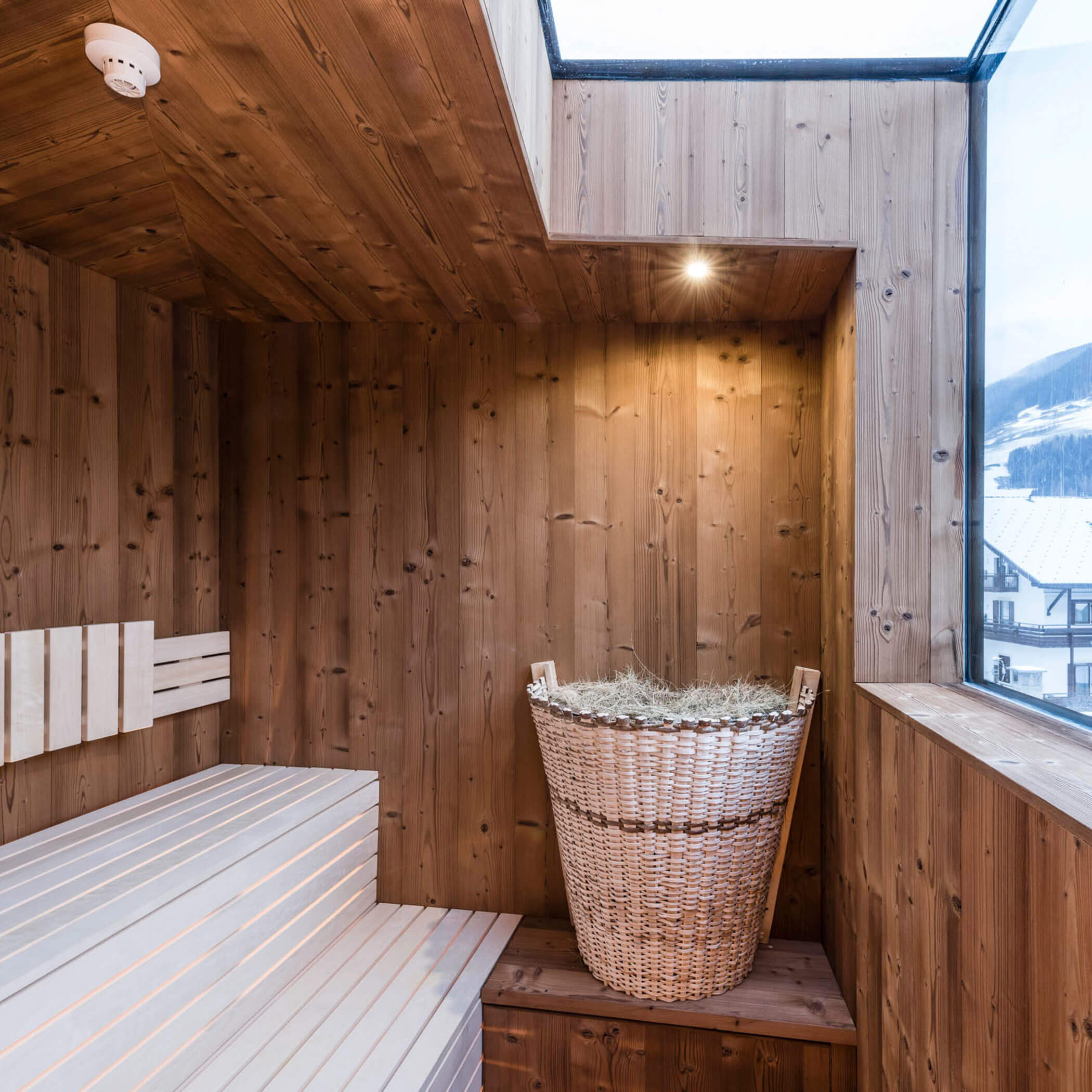Modern wooden sauna with a basket of birch twigs and a large window overlooking snowy buildings outside.