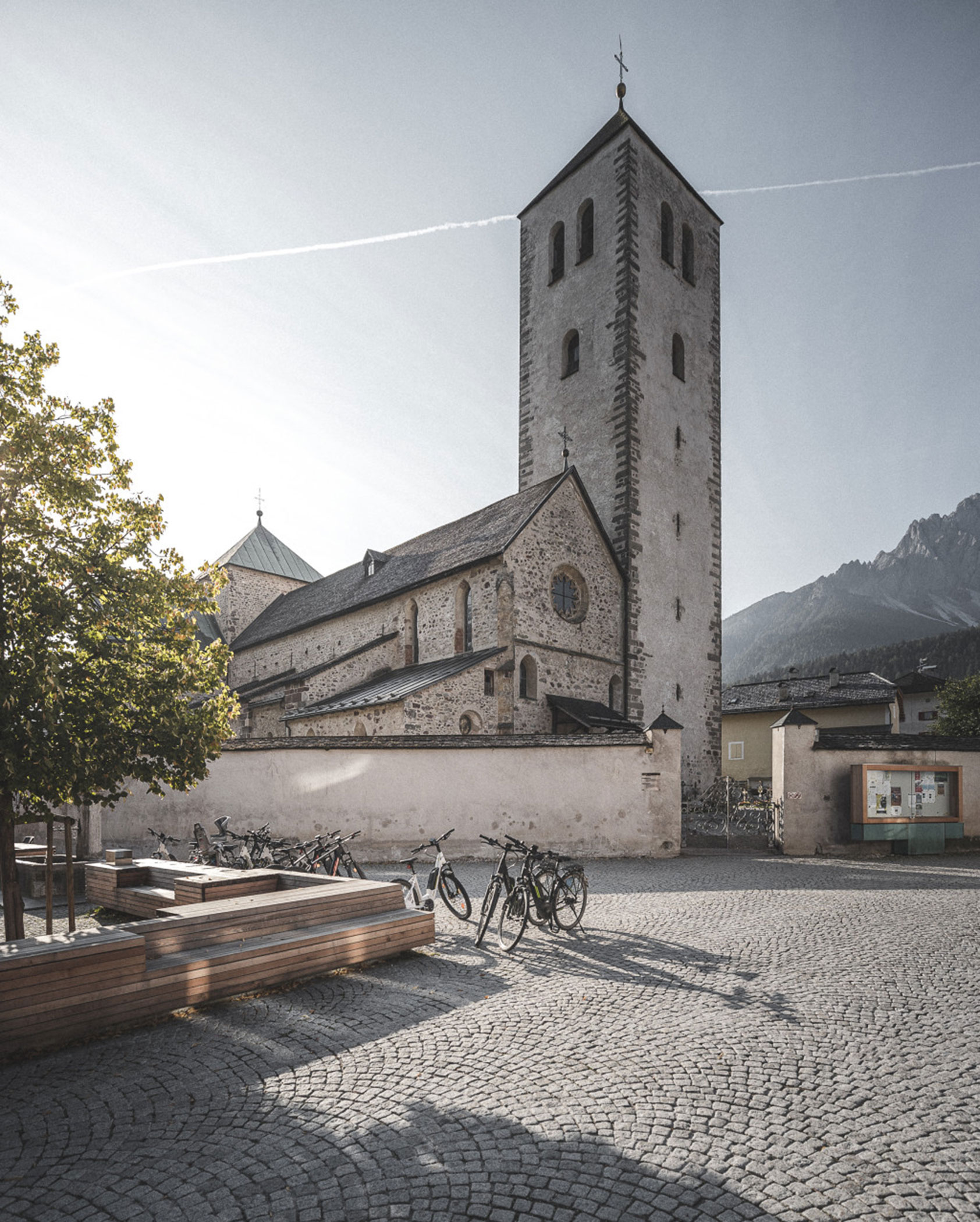 A stone church with a tall tower, bicycles, and benches in a sunlit cobblestone square.