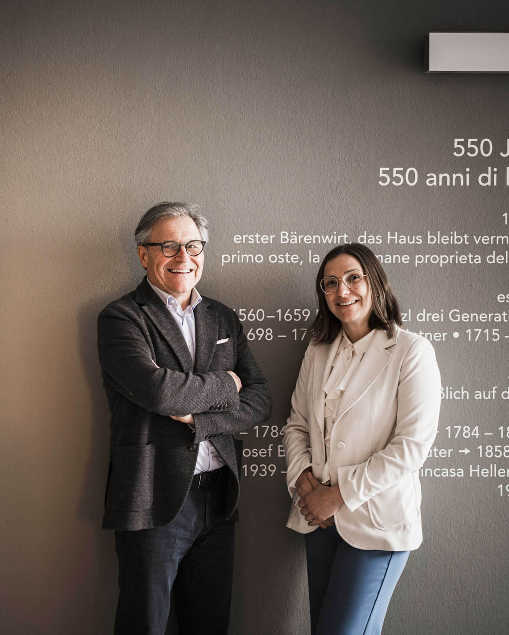 Two people in business attire smiling and standing in front of a wall with historical text in multiple languages.