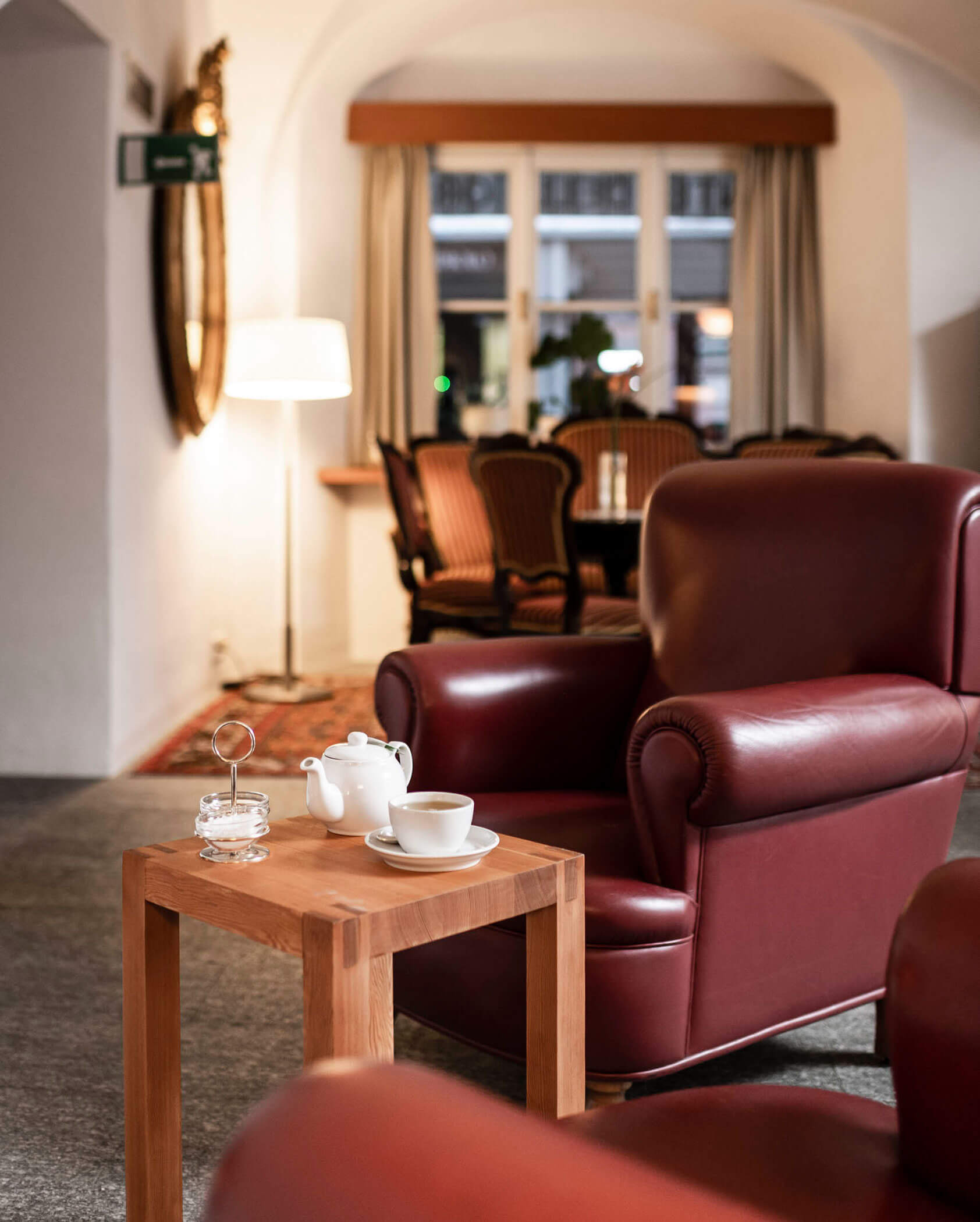 A maroon leather armchair by a wooden table with a teapot and cup in a warmly lit lounge.