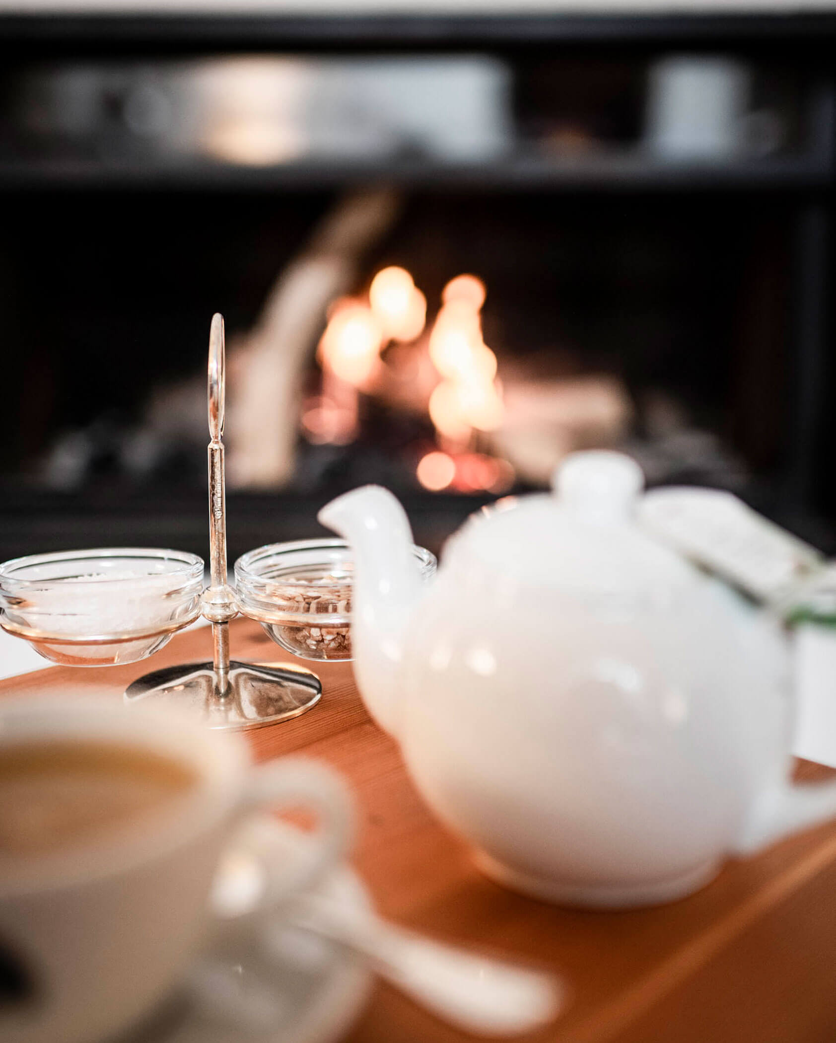 A teapot, teacup, and glass dishes on a table with a fireplace glowing in the background.