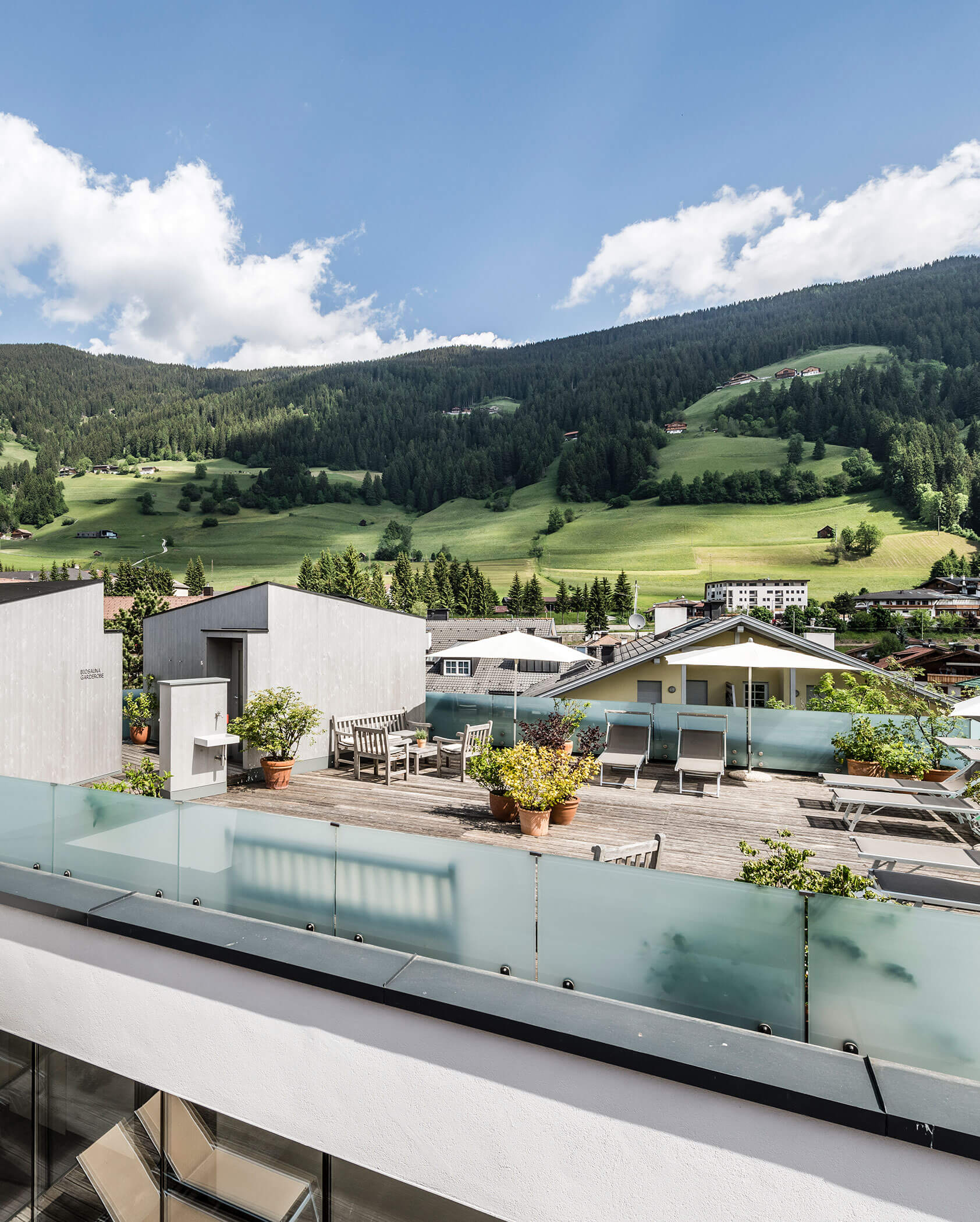 A rooftop terrace with lounge chairs and potted plants, overlooking green hills and mountains under a blue sky.