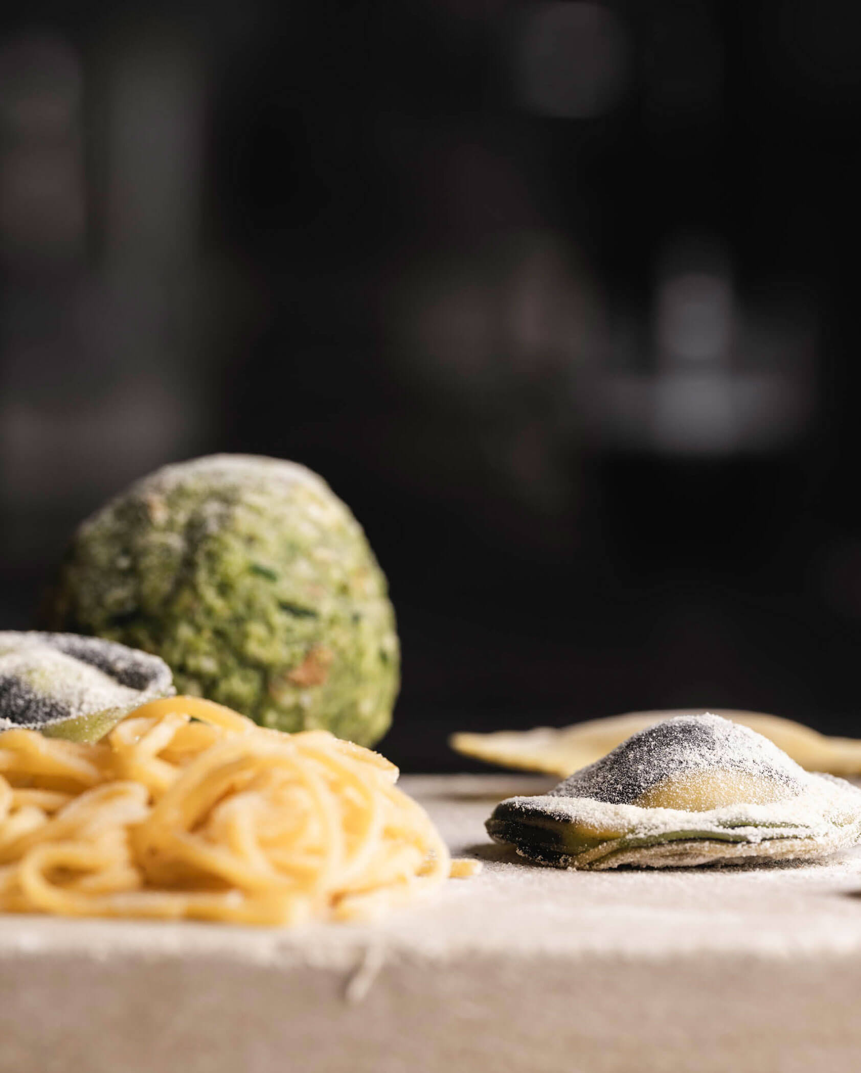 Fresh pasta, including noodles, stuffed ravioli, and a ball of green dough, dusted with flour on a surface.