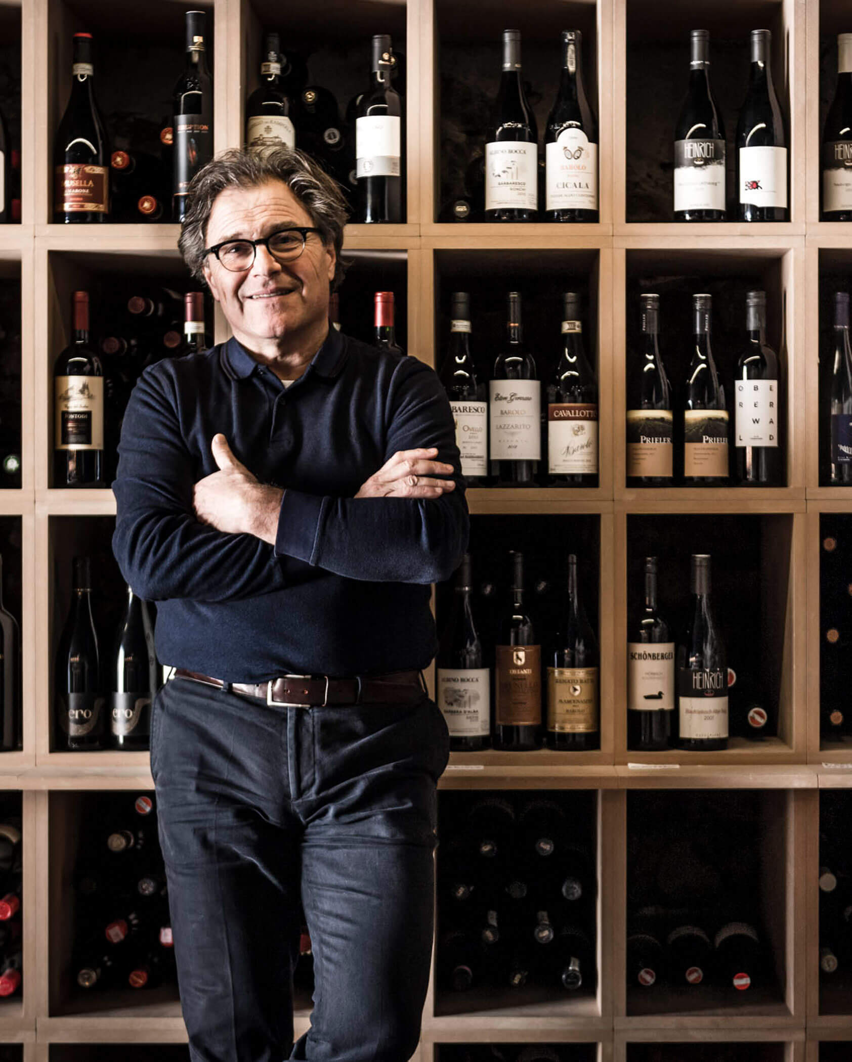 Smiling man in dark clothes stands in front of a wall of wine bottles displayed on wooden shelves.