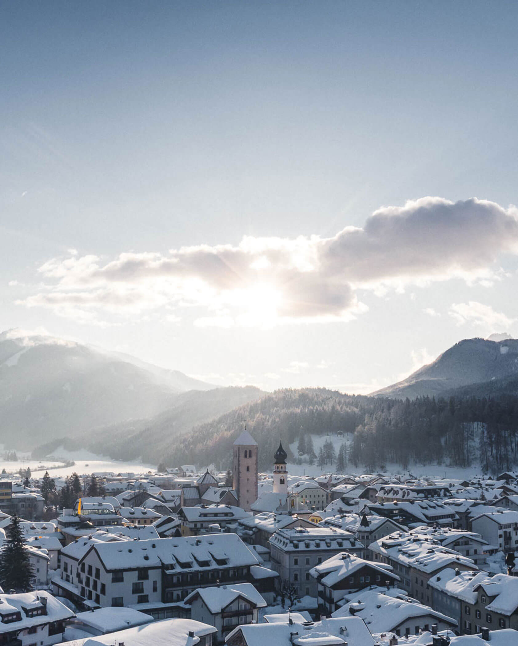 Snow-covered town with church towers, surrounded by mountains under a bright, partly cloudy sky at sunrise.