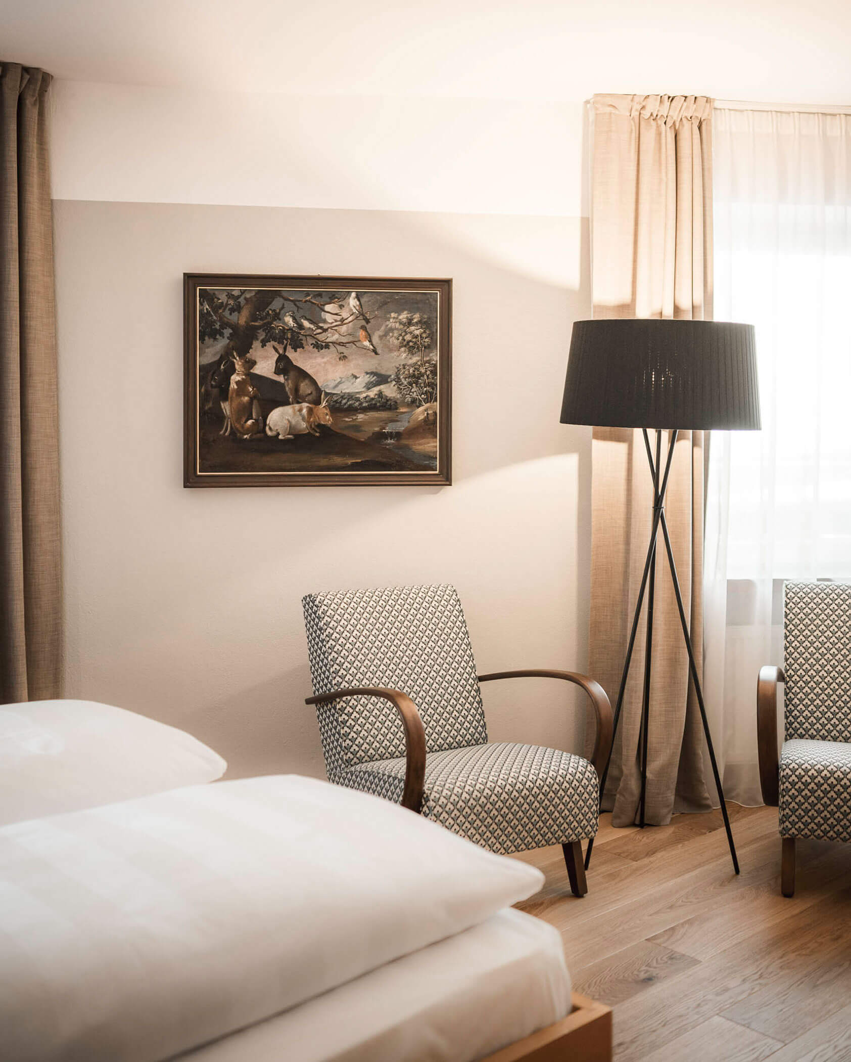 A cozy bedroom with two patterned armchairs, a floor lamp, a bed, curtains, and a framed painting on the wall.