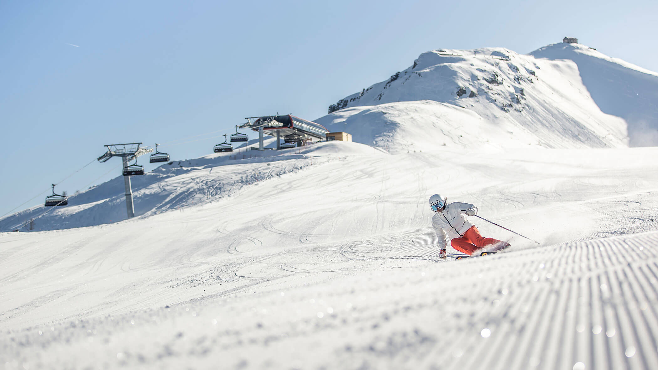 A skier in red pants carves down a snowy slope with ski lifts and a mountain in the background.