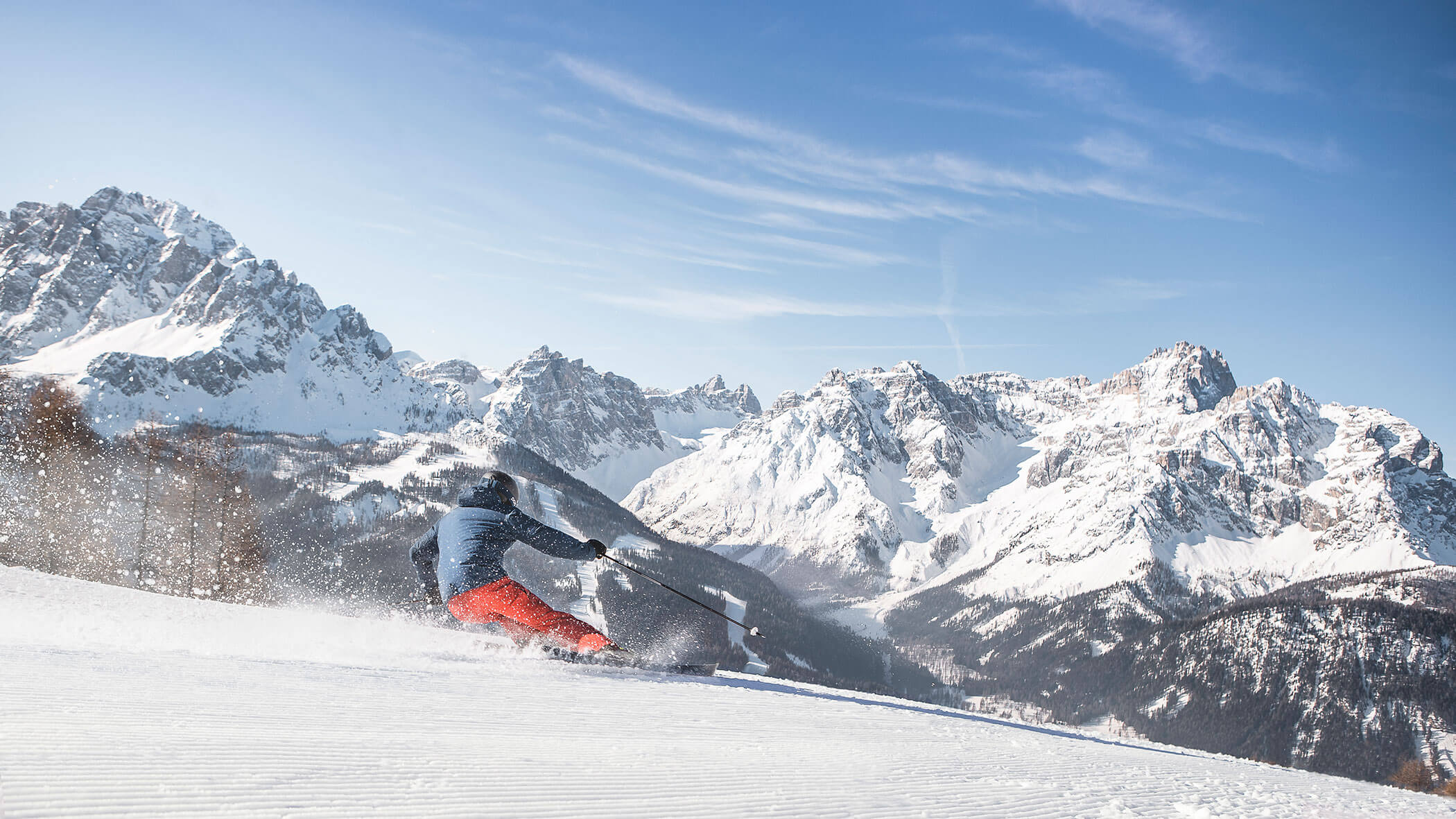 A skier in red pants skis down a snowy slope with tall, snow-covered mountains in the background.