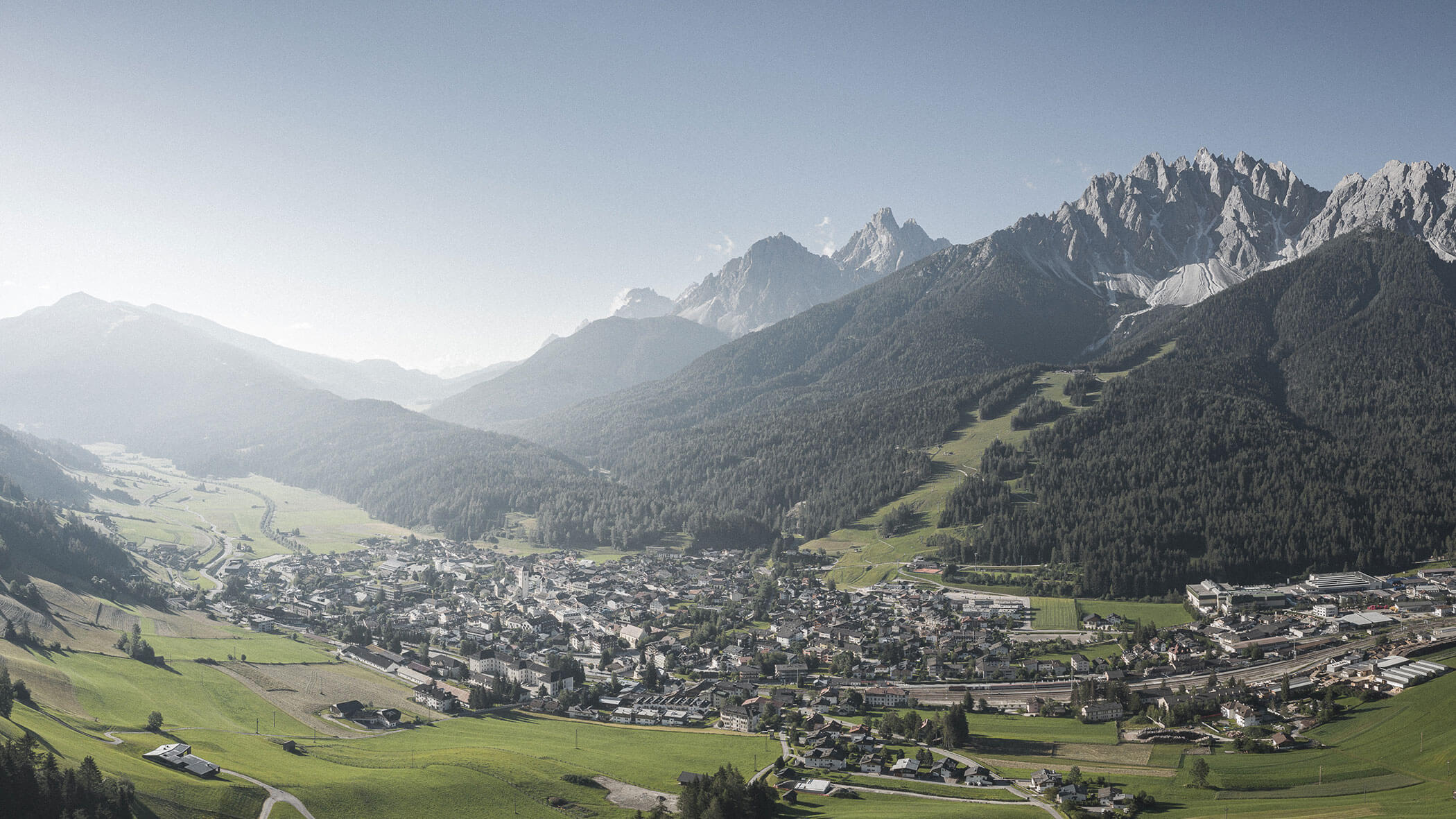 Una città immersa in una valle verde circondata da montagne boscose e picchi rocciosi sotto un cielo limpido.