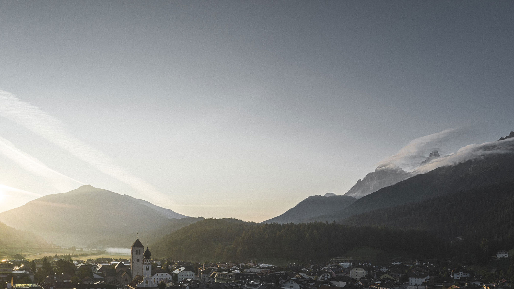 Vista panoramica di un villaggio di montagna all'alba, con colline nebbiose e il campanile di una chiesa in primo piano.