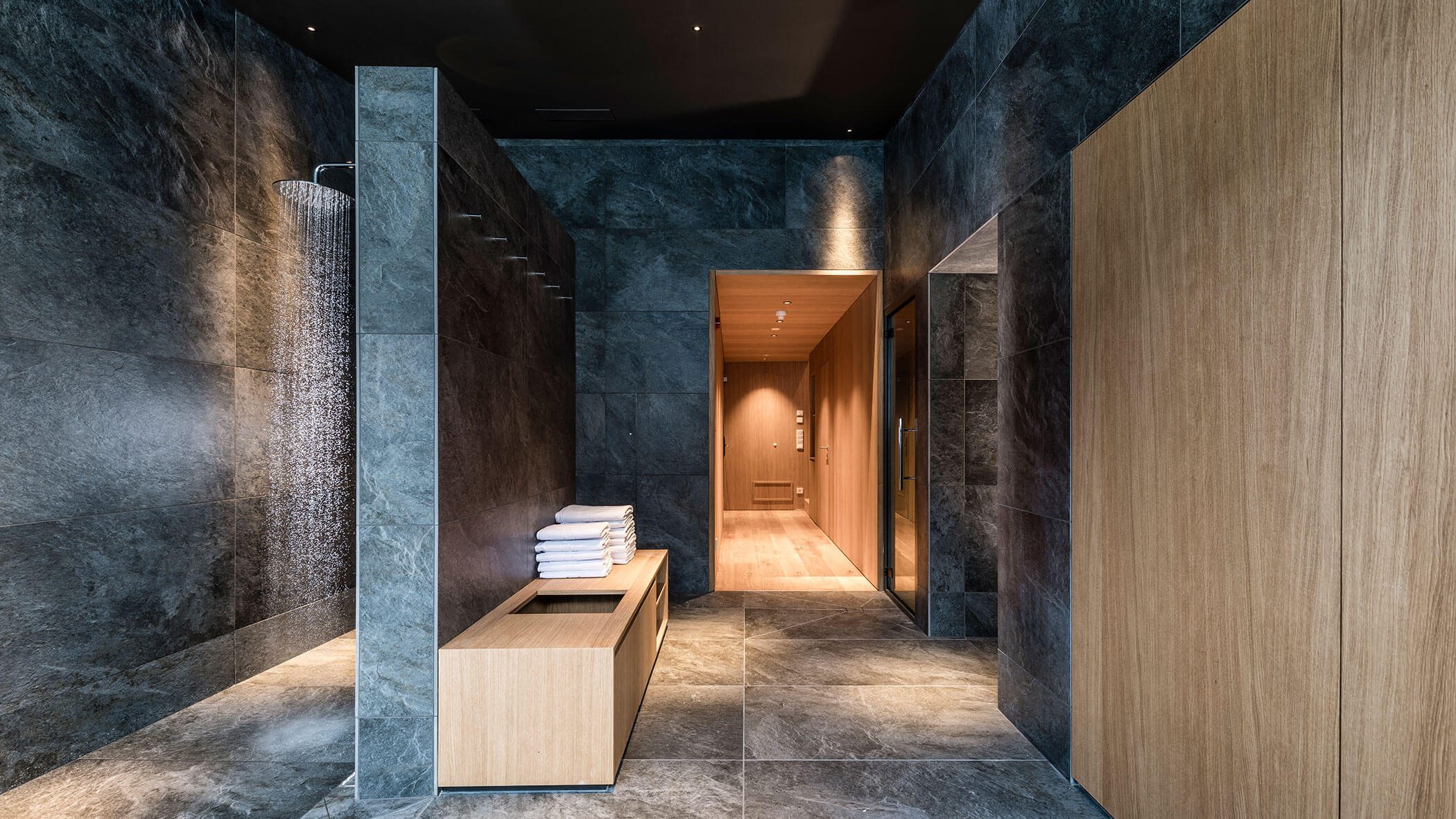 Modern, minimalist shower room with dark stone tiles, wooden accents, a bench, and neatly stacked white towels.