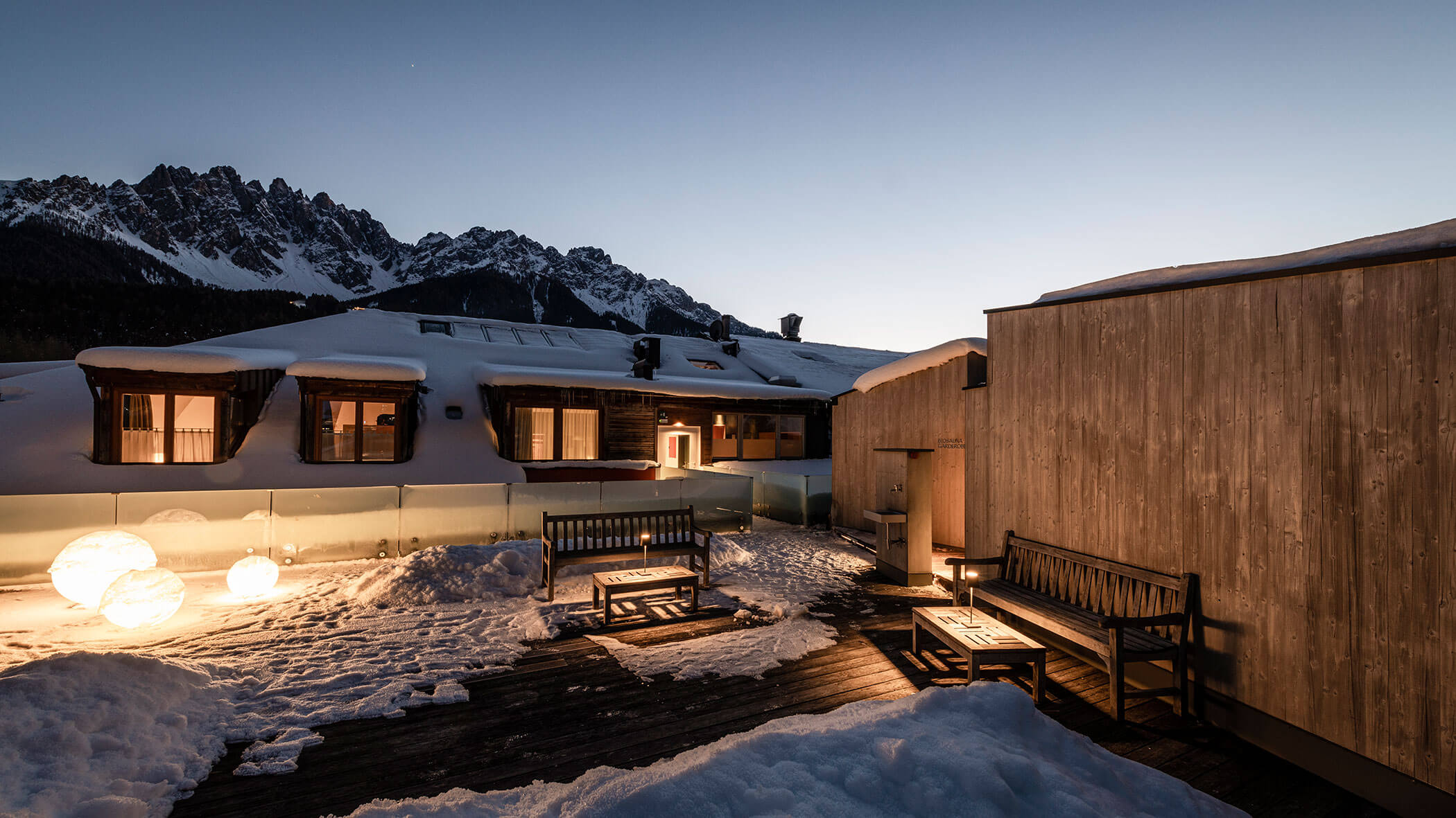 Terrazza innevata sul tetto con panchine, luci scintillanti e cime di montagna sullo sfondo al tramonto.