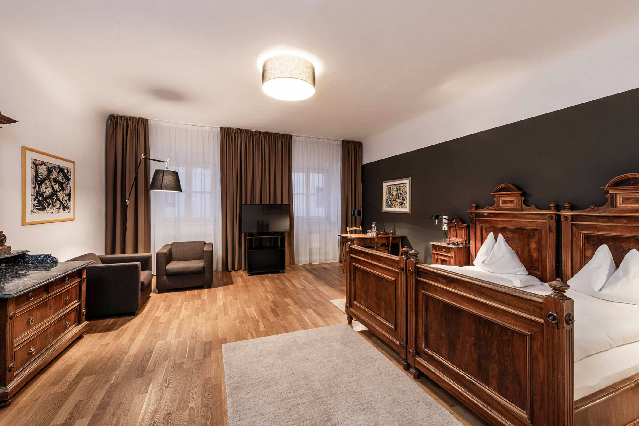 Spacious hotel room with antique wooden beds, seating area, TV, and large windows with brown curtains.