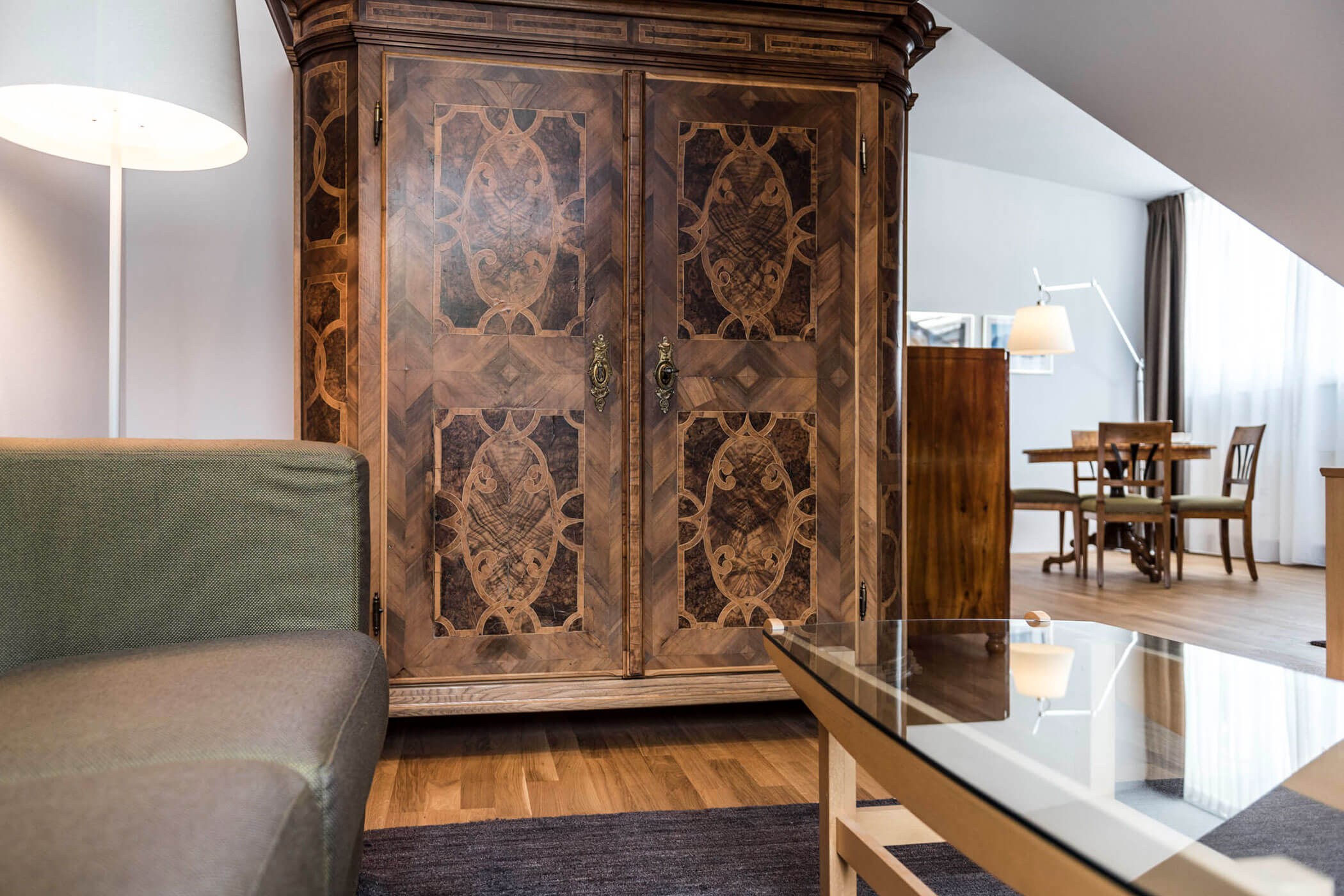 Large ornate wooden wardrobe beside a sofa and glass coffee table in a bright, modern living room.