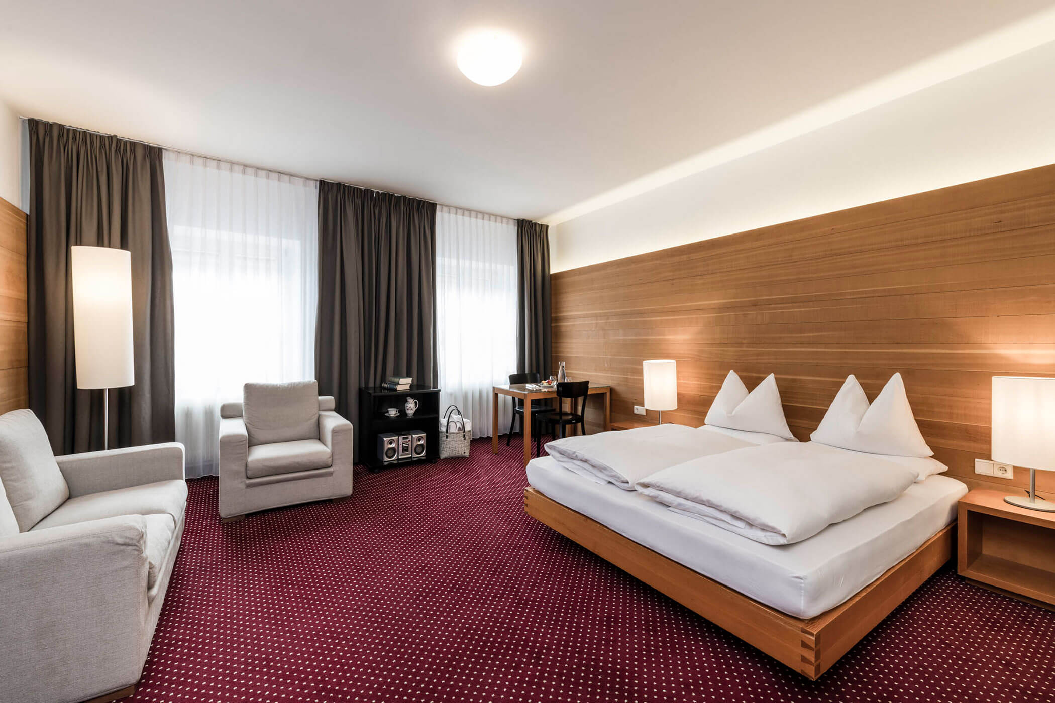 Modern hotel room with a double bed, sofa, armchair, desk, and large windows with dark curtains.