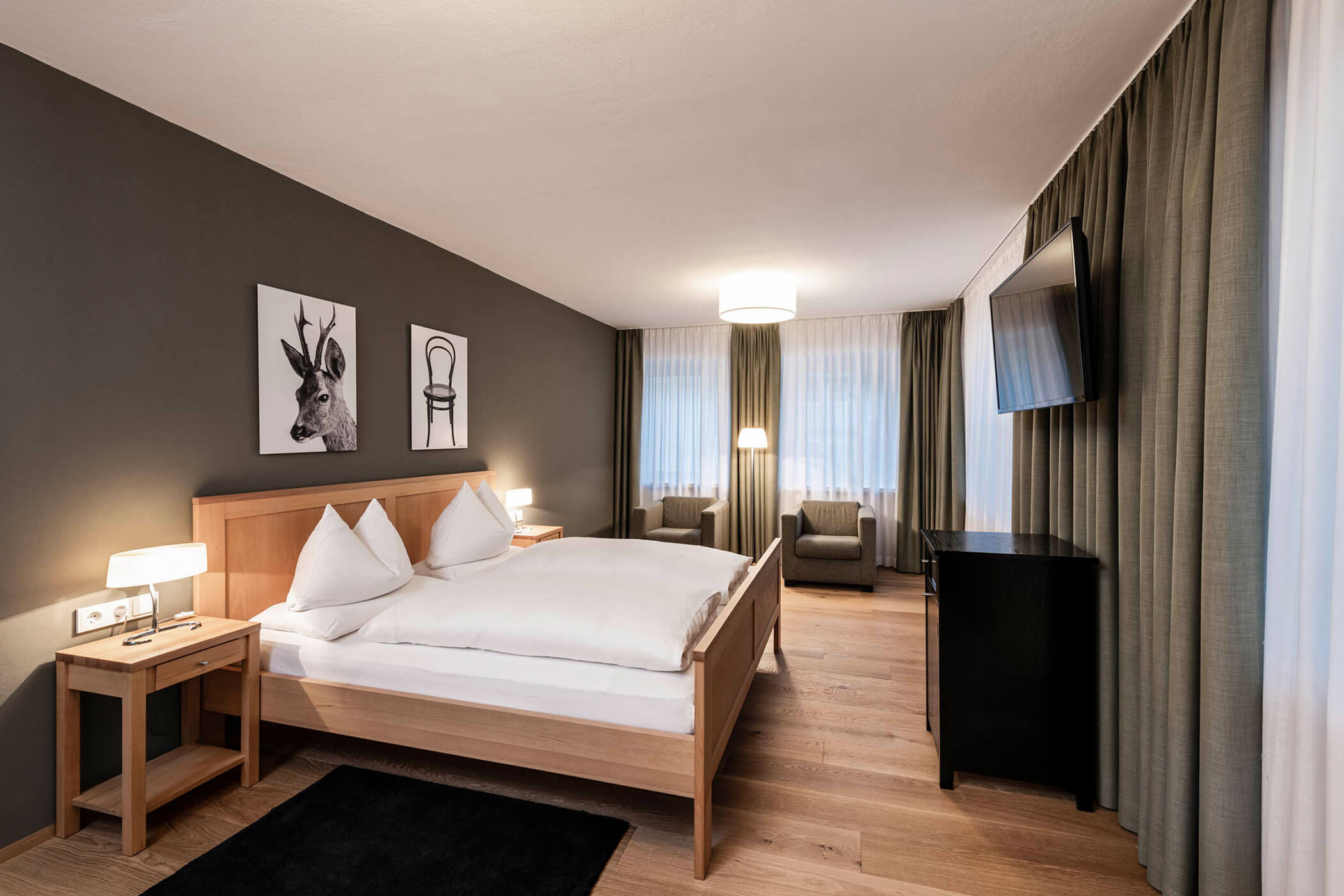 Modern hotel room with double bed, artwork, armchair by window, TV, and warm lighting.