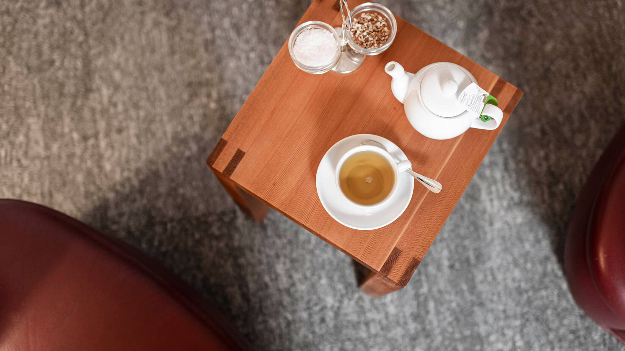 White teacup, teapot, and sugar bowls on a small wooden table with gray carpet and red chairs nearby.