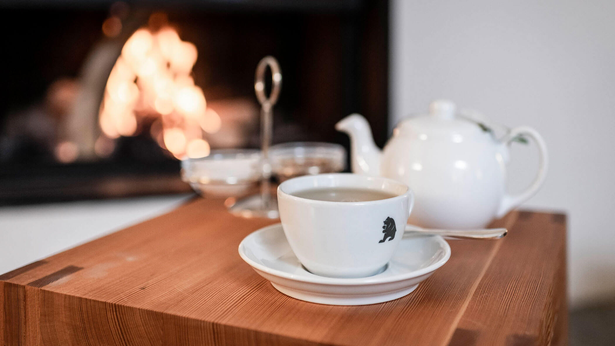 A white teacup and saucer sit on a wooden table, with a teapot and fire burning in the background.