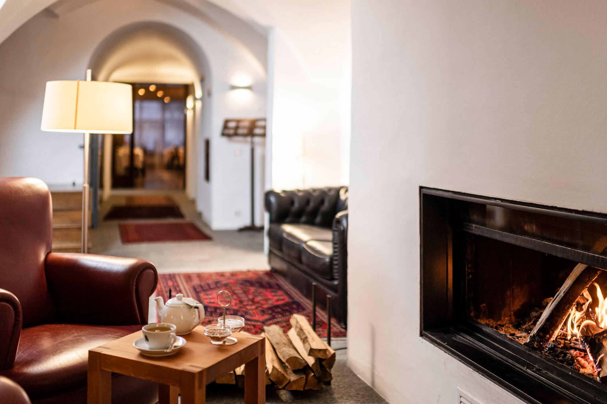 Cozy lounge with armchair, coffee table, teapot, fireplace, and sofas in a warmly lit room.